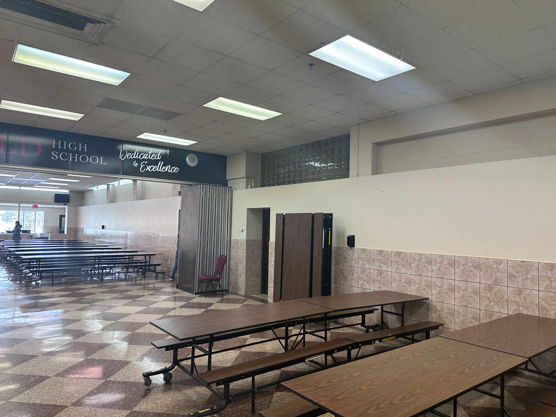 Empty school cafeteria with brown tables, checkered floor, and off-white walls.