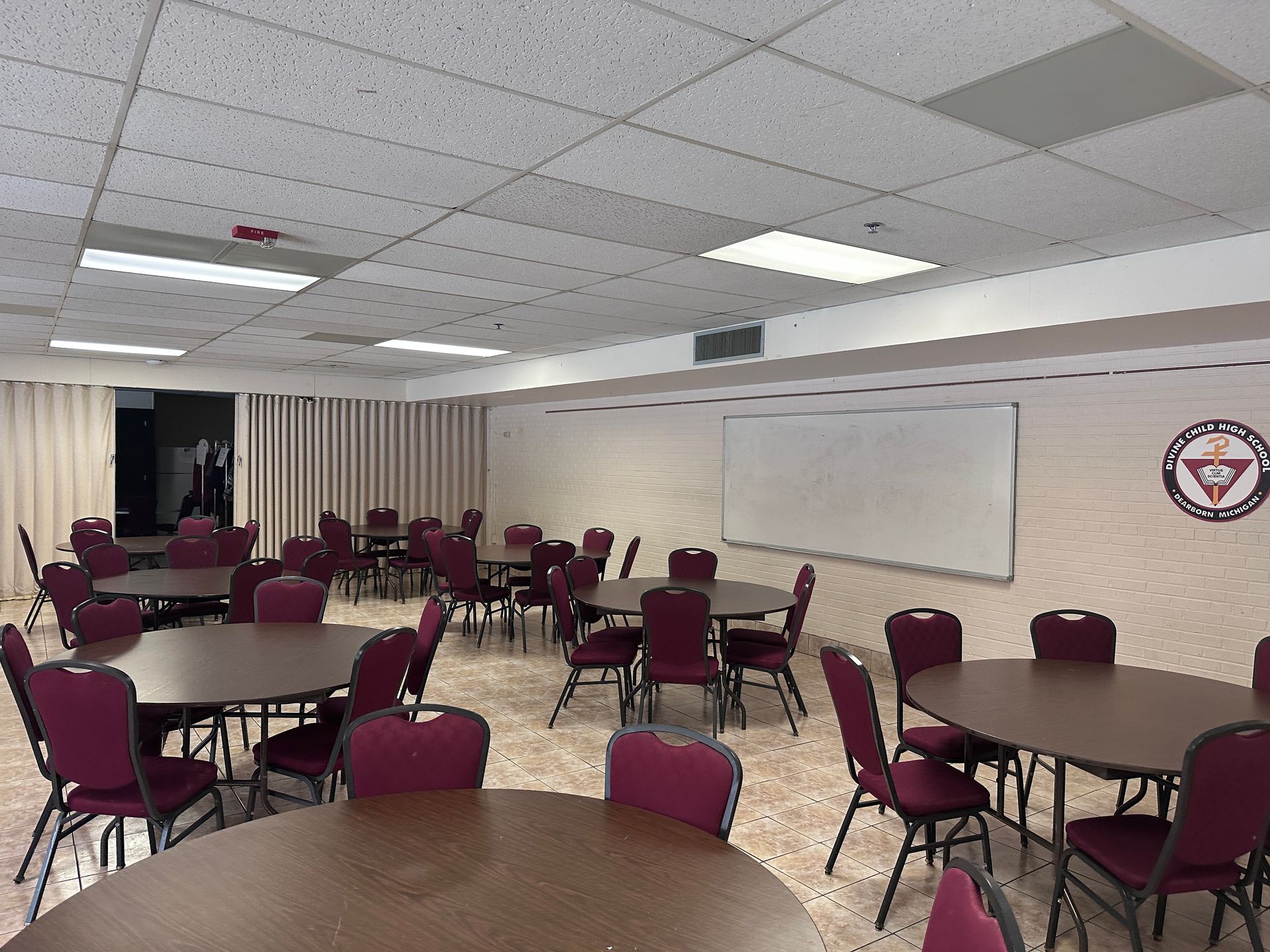 A room with round tables and maroon chairs. A whiteboard and a logo are on the wall.