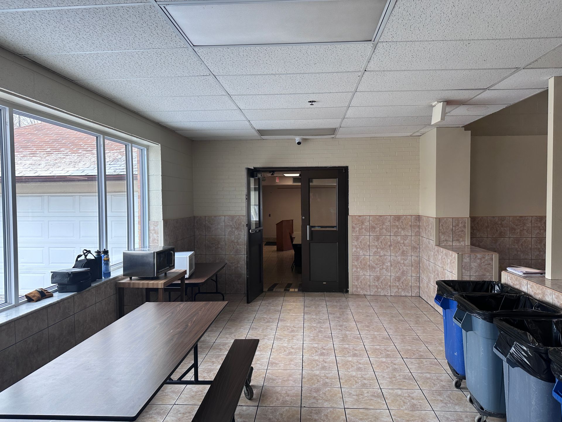 Empty cafeteria with tables, windows, a doorway, trash cans, and tiled walls.