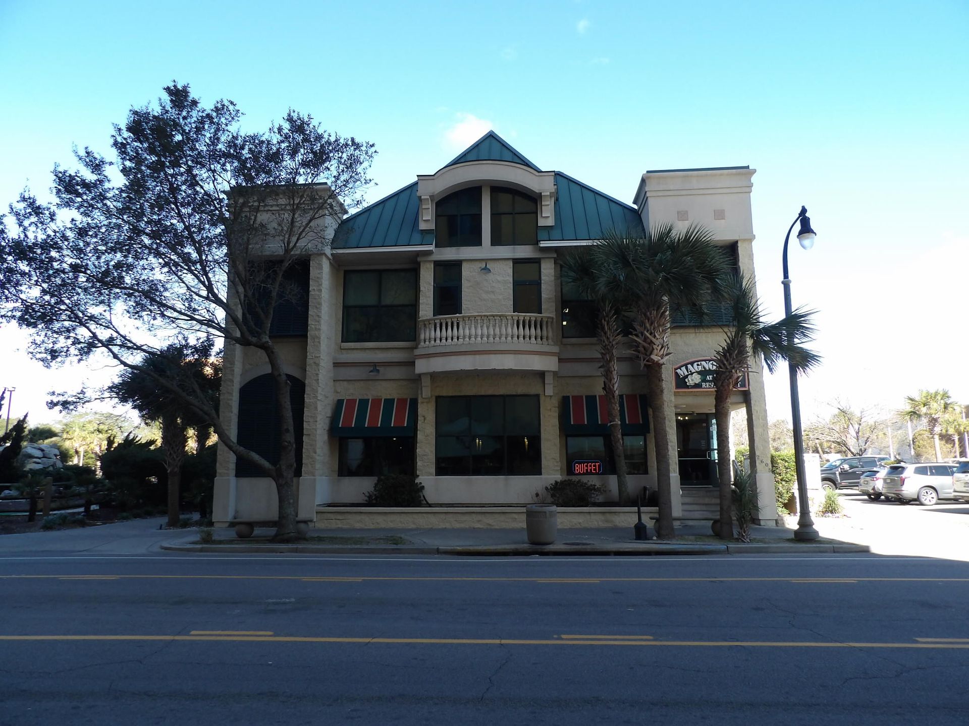 Two-story beige building with teal roof and balcony, palm trees and street in front.