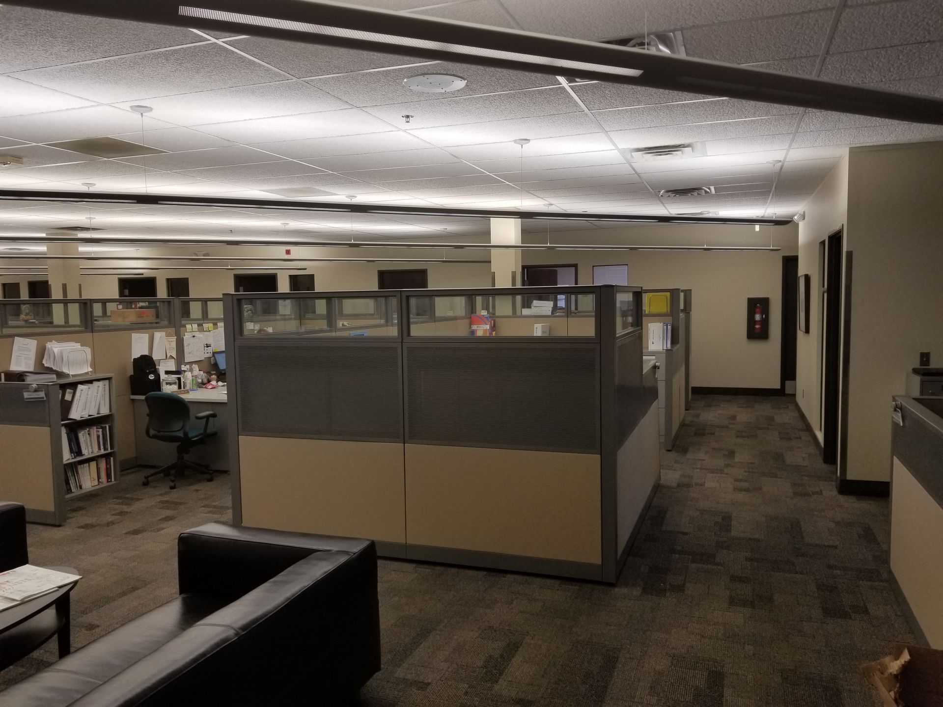 Office cubicles in a low-lit room with a sofa in the foreground and a hallway in the background.