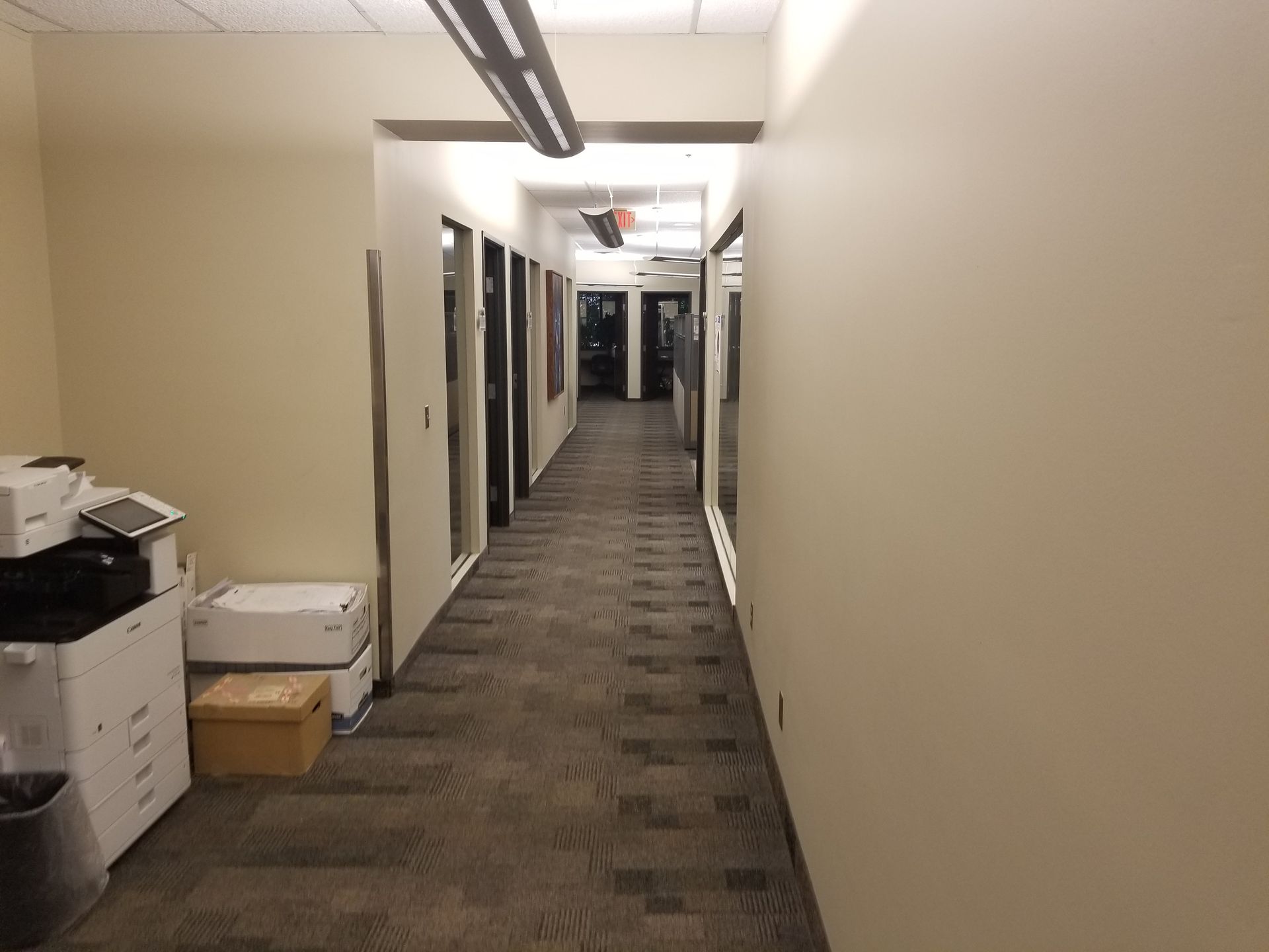 Long hallway with doors on the left and beige walls. Printers and boxes are on the left.
