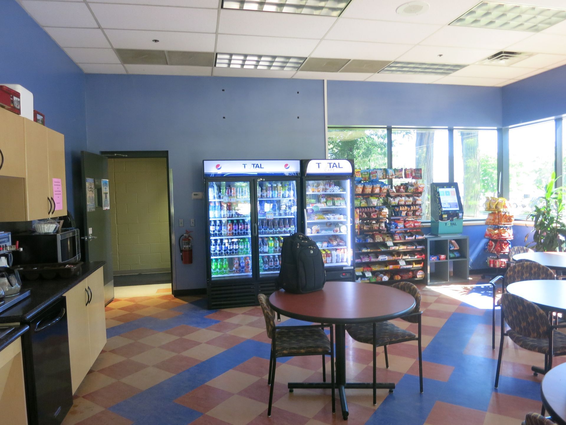 Cafeteria with beverage coolers, snacks, tables, chairs, and a kitchen area; blue and red checkered floor.