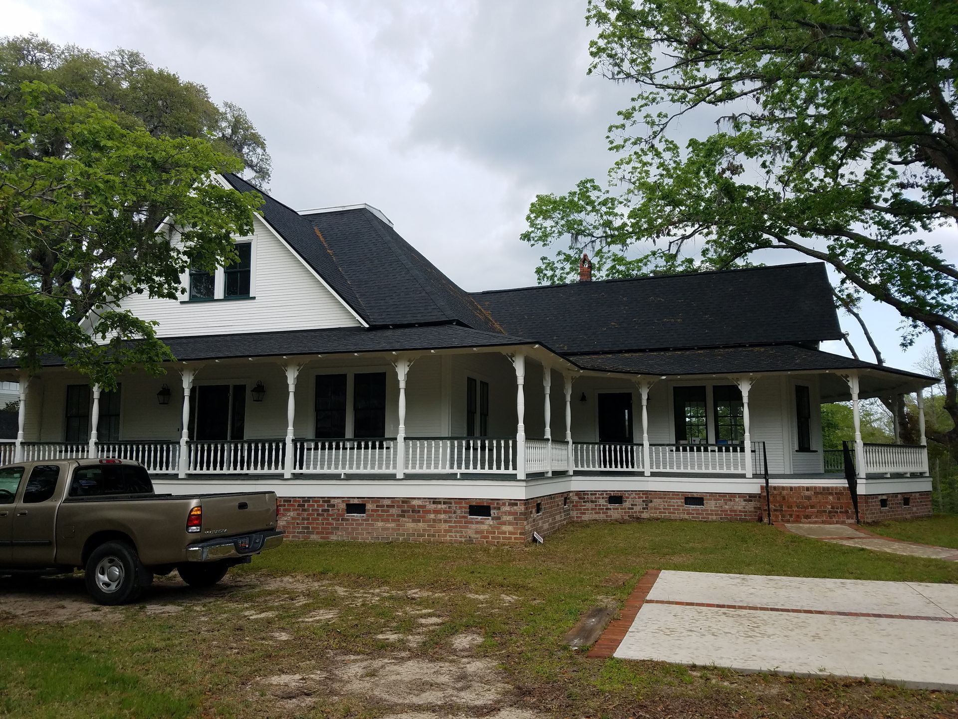 White farmhouse with wrap-around porch, brown truck parked in front, overcast sky.