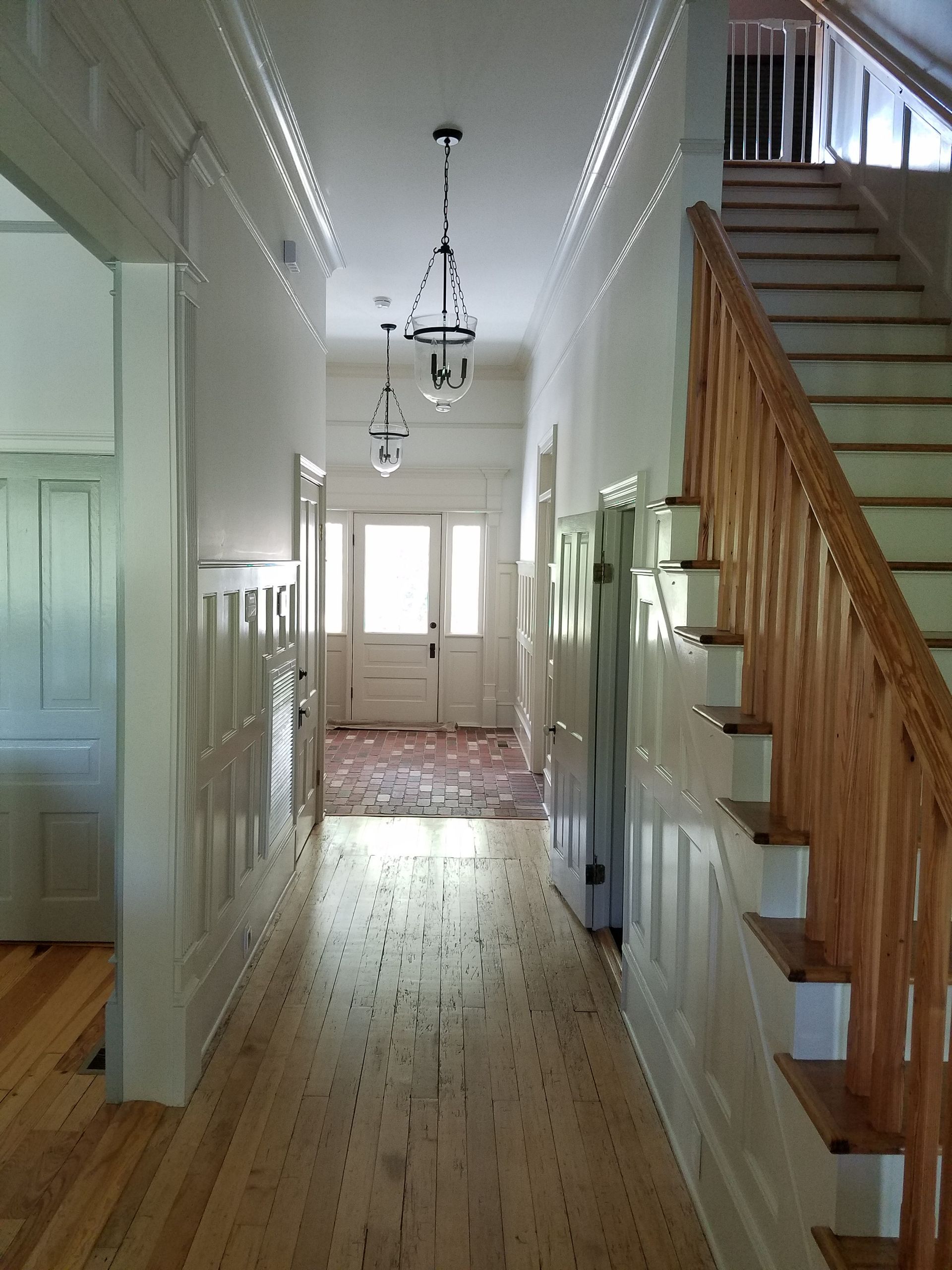 Long hallway with wood floors, staircase, and entry door at the end. White walls with decorative trim and hanging lights.
