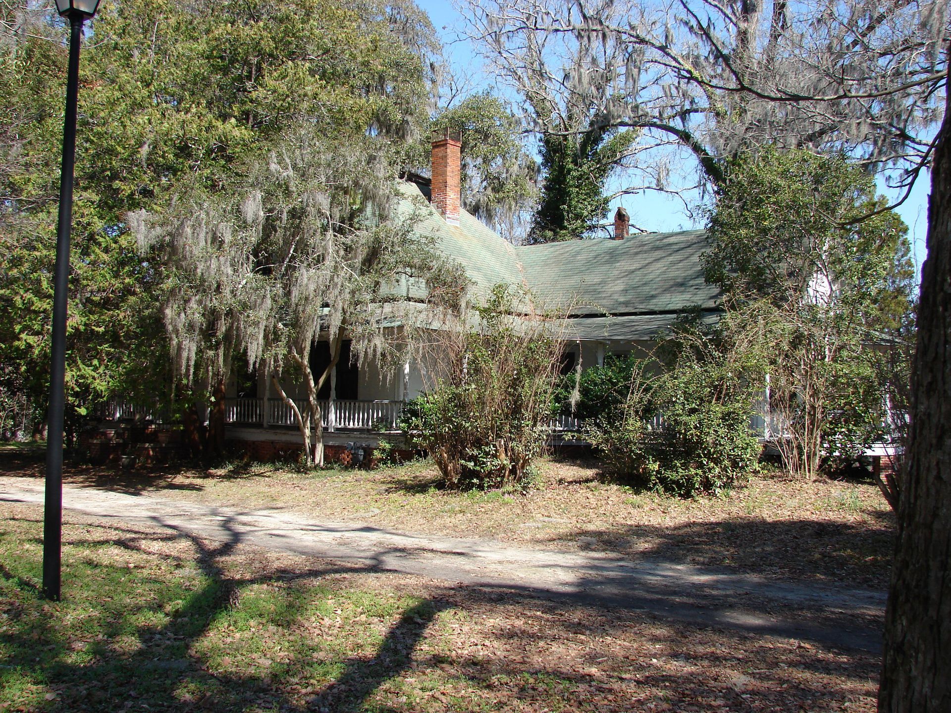 Old white house with green roof, overgrown with trees and plants, under a blue sky.