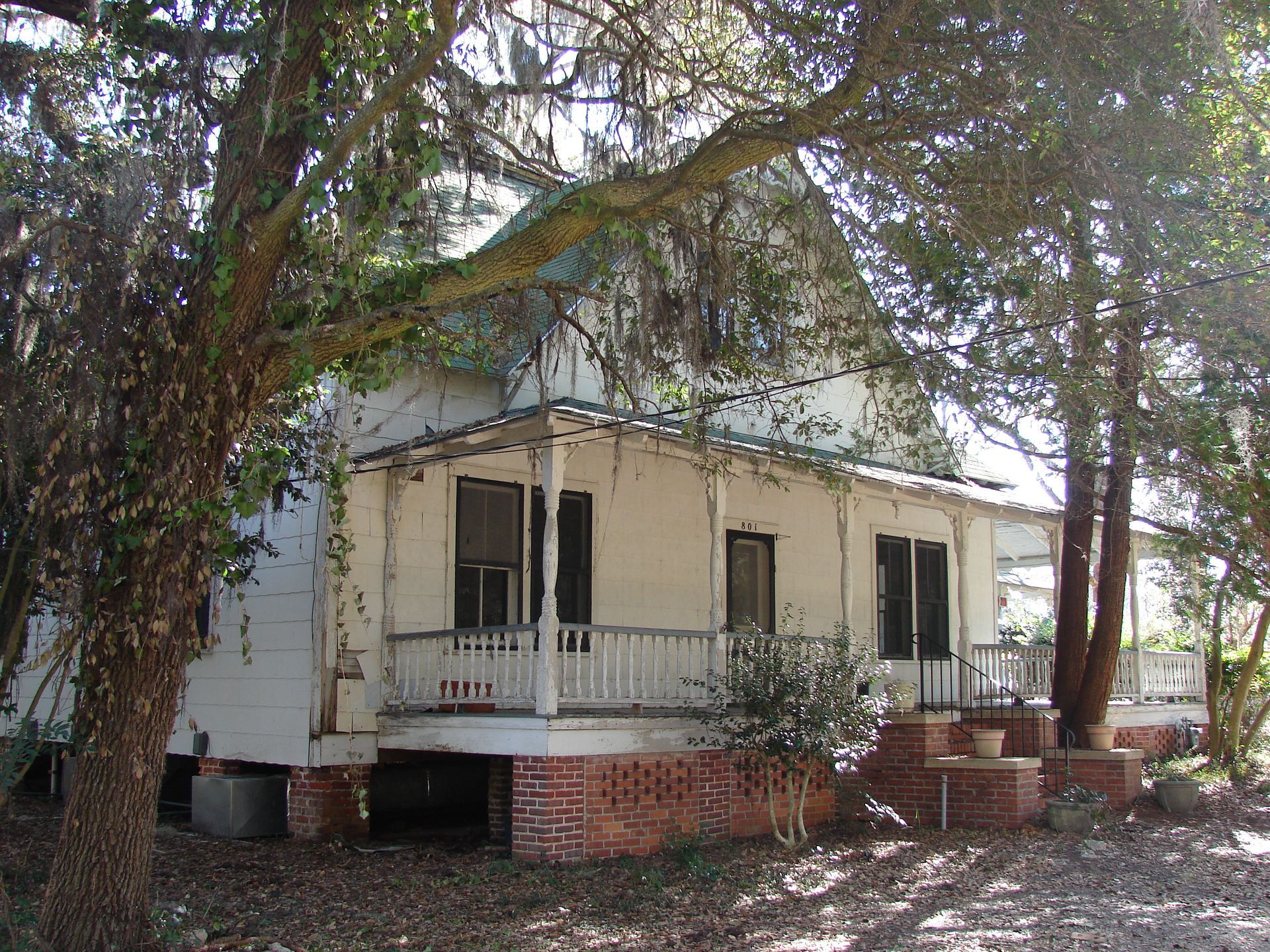 Dilapidated white house with a porch, surrounded by trees.