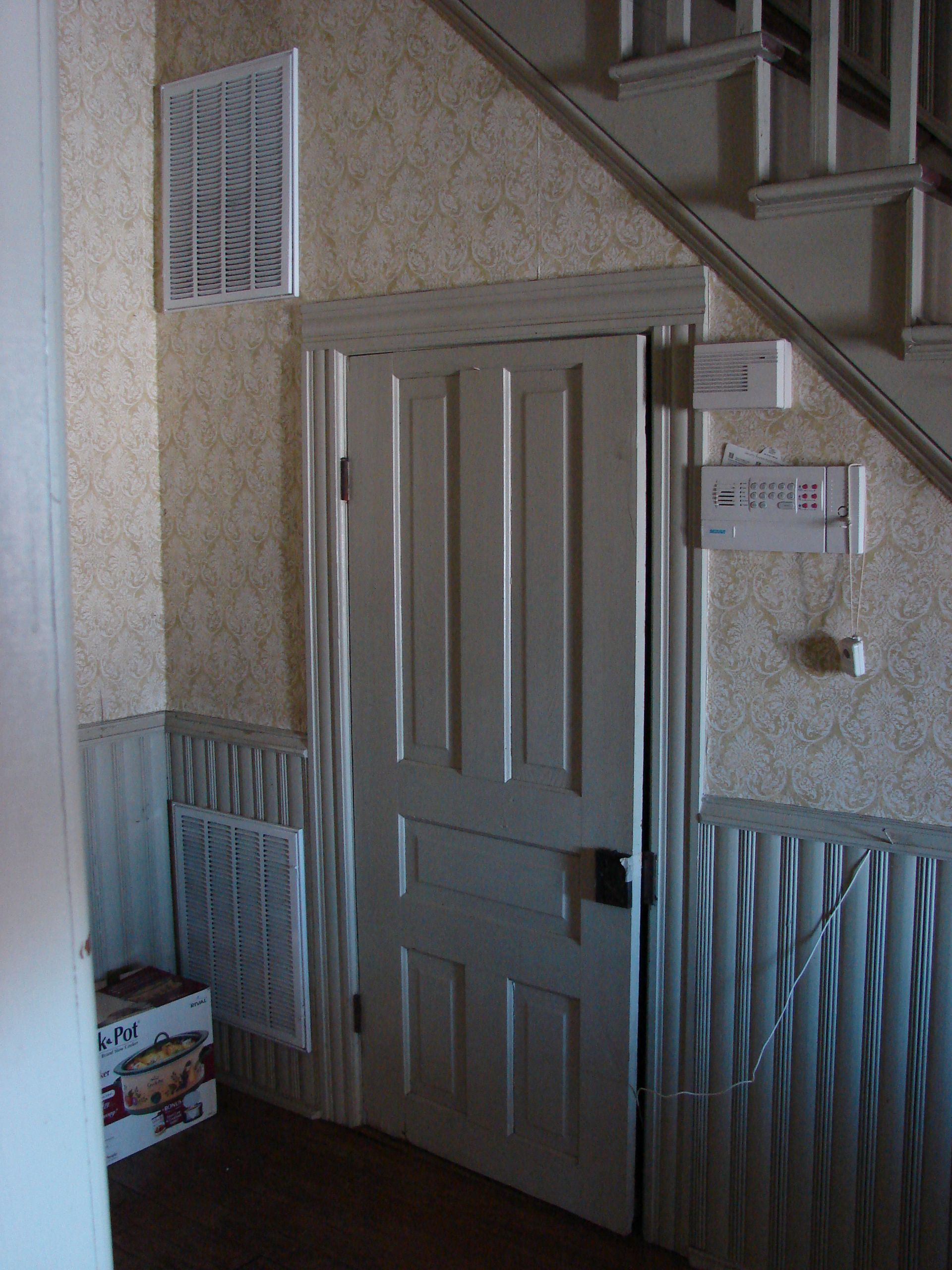 Door under a staircase, near an air vent and radiator. Light interior, wallpaper, and a box on the floor.