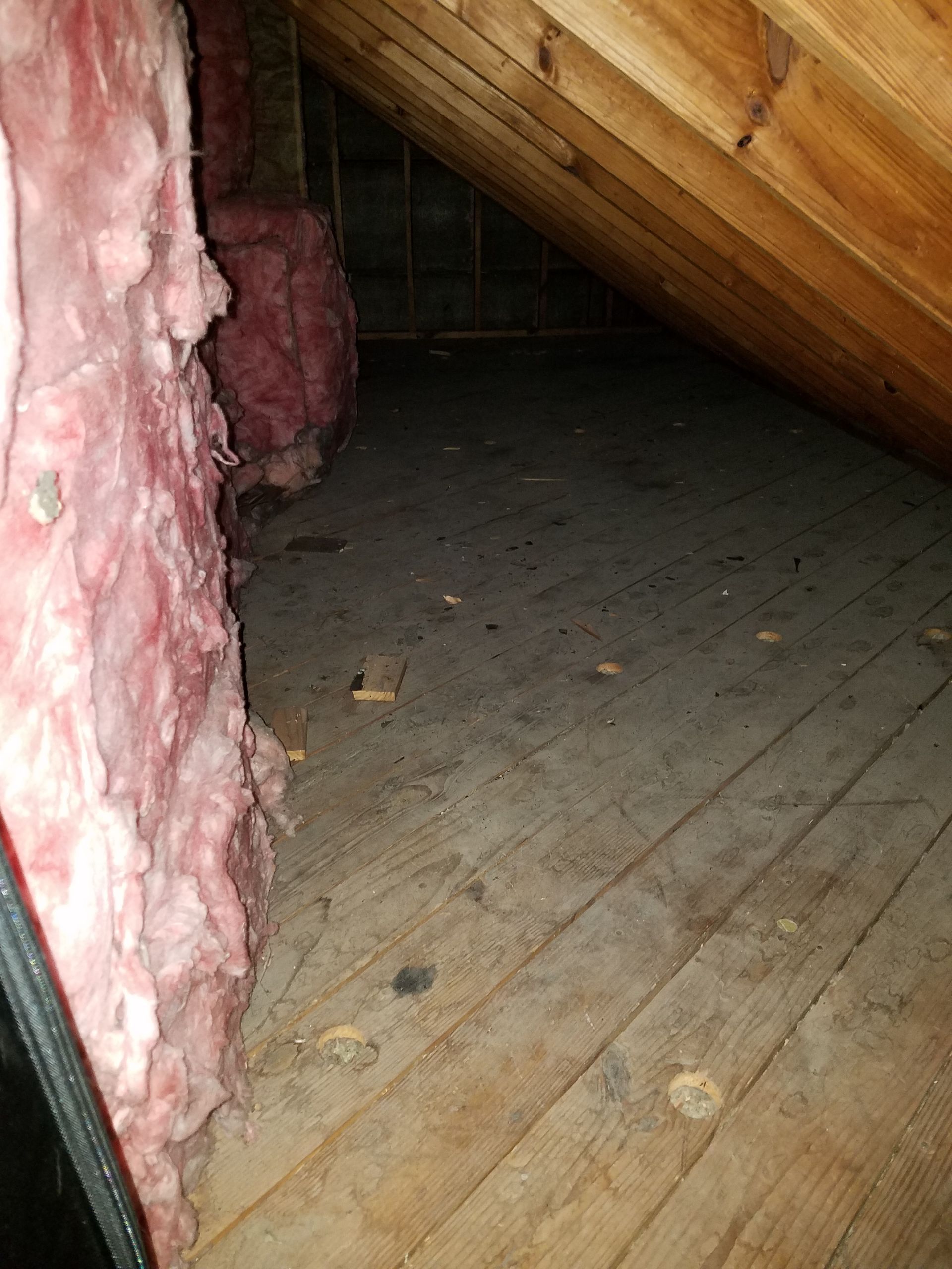 View inside an attic. Pink insulation is visible near a wooden floor. Wooden rafters are above.