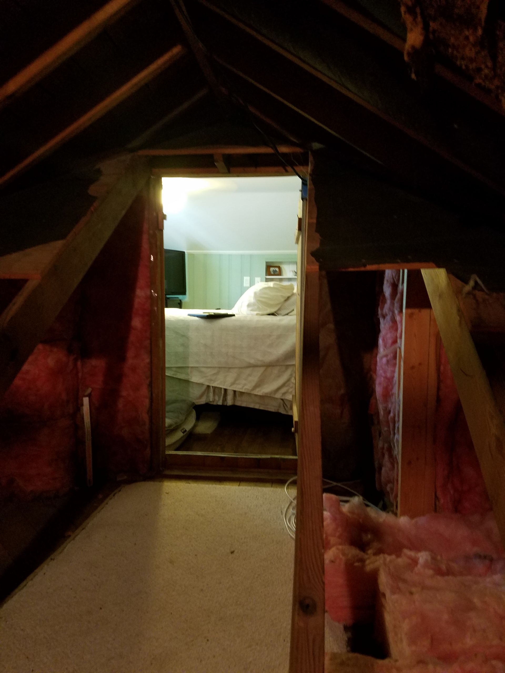 Looking from attic, doorway reveals bedroom with white bed, walls are pink insulation and bare wood beams.