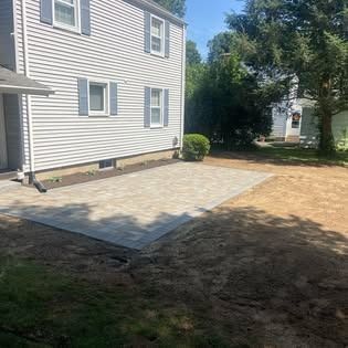 Backyard with new gray paver patio adjacent to a white house with blue shutters.