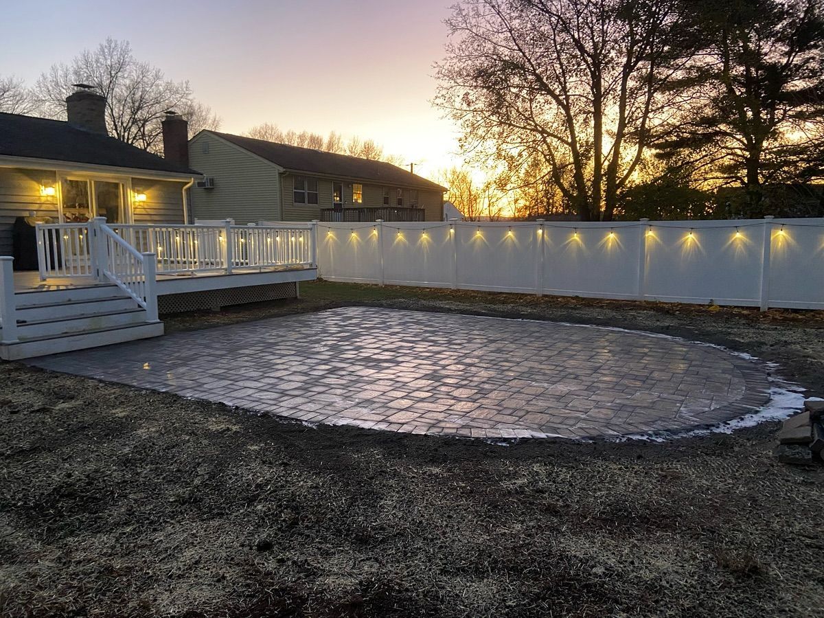 Backyard with deck, oval-shaped gravel area, white fence with lights, and sunset sky.