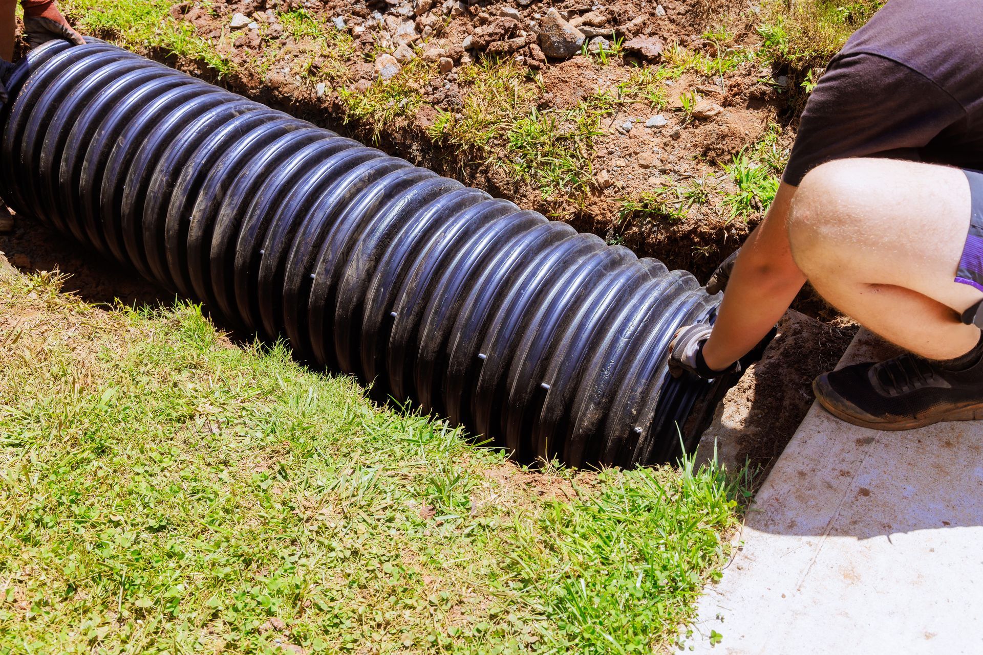 Black corrugated drain pipe being installed beside a grassy yard, with a person kneeling nearby.
