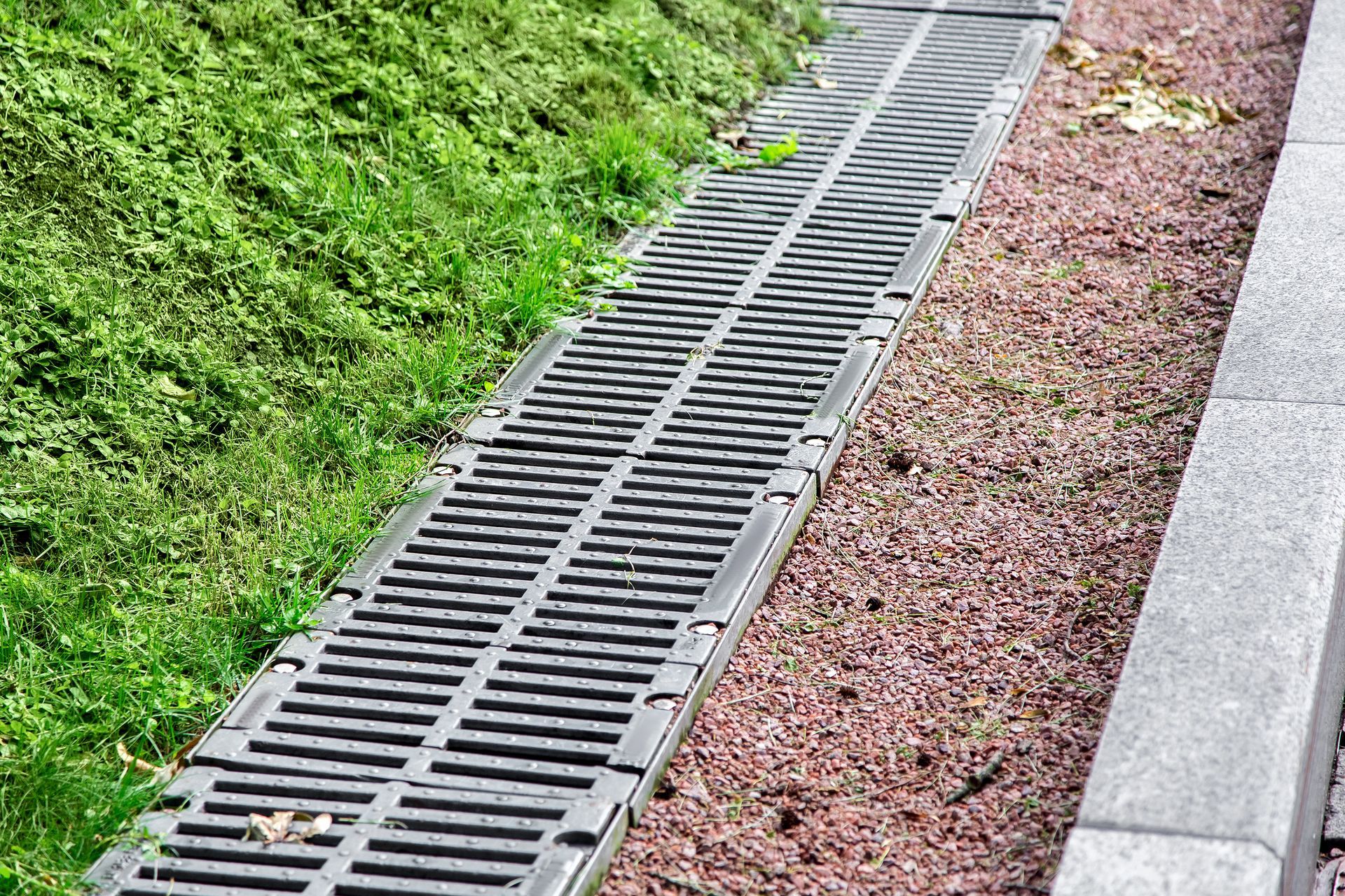 Storm drain grate beside a grassy slope and gravel strip next to a concrete path