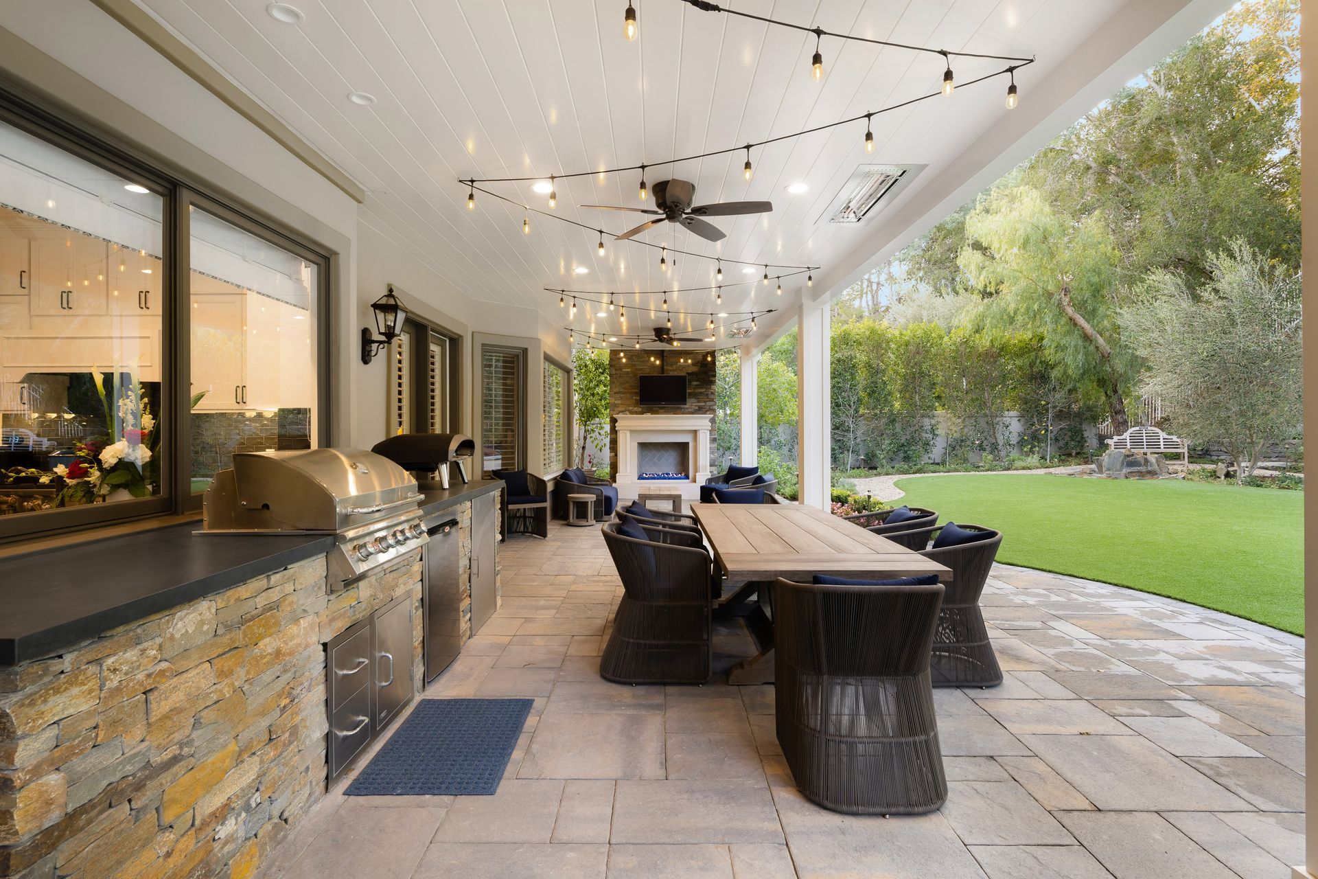 Outdoor patio with grill, fireplace, dining table, and string lights, overlooking a grassy yard.