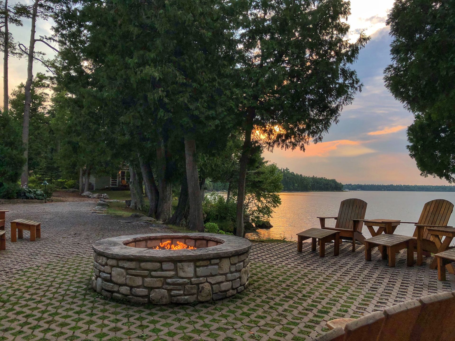 Lakeside fire pit with Adirondack chairs on a stone patio at sunset, trees and calm water in the background