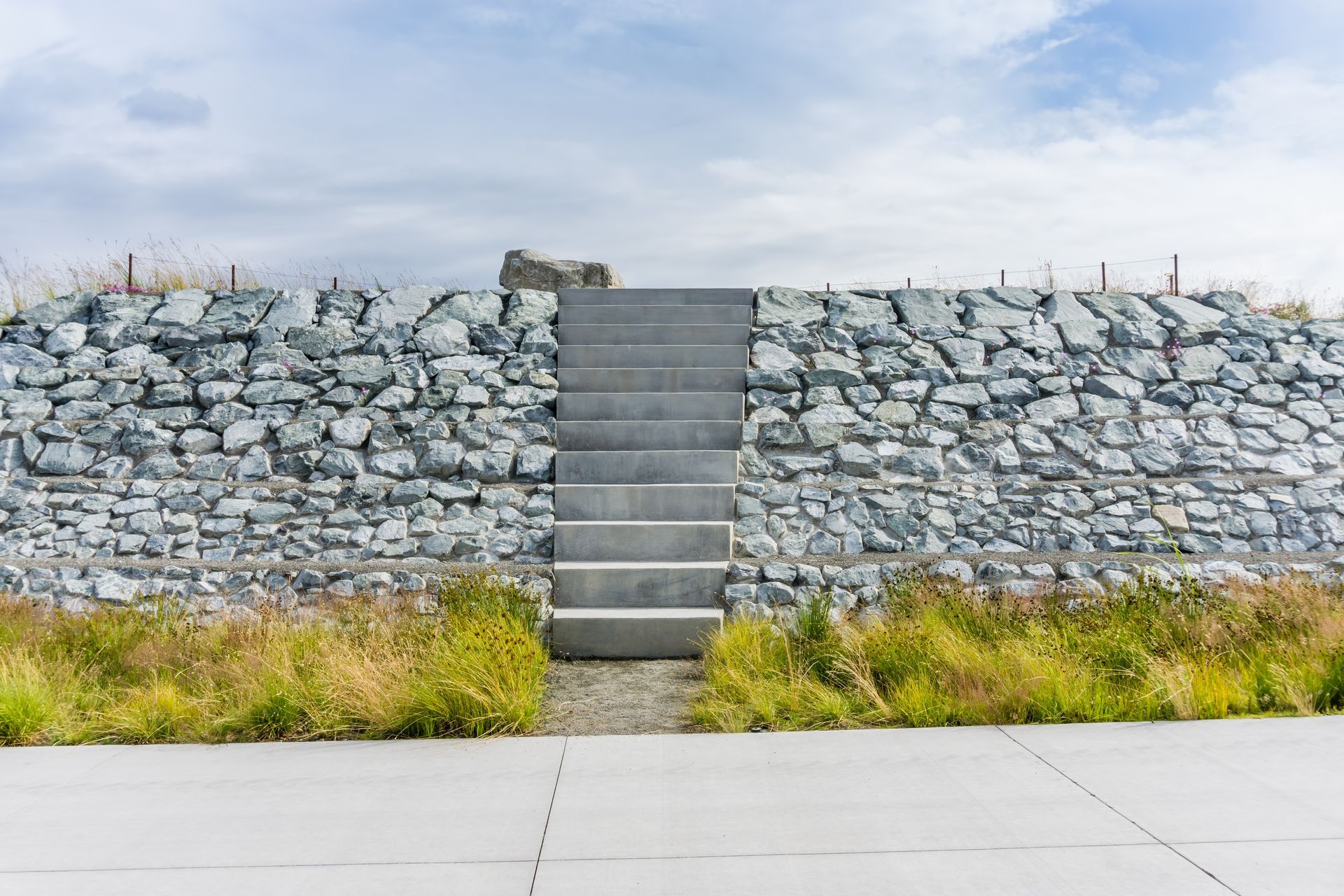 Stone retaining wall with central concrete stairs above a sidewalk and patches of grass