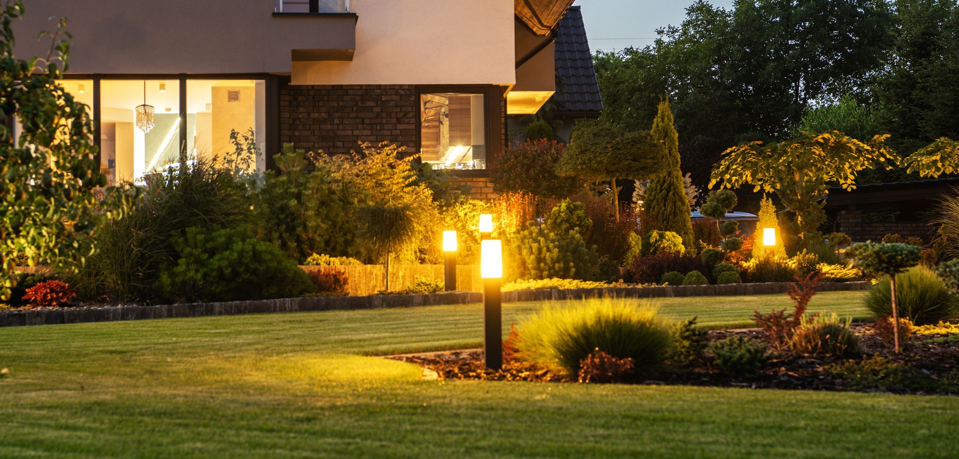 Modern house at dusk with a lit garden, lawn, shrubs, and pathway lights