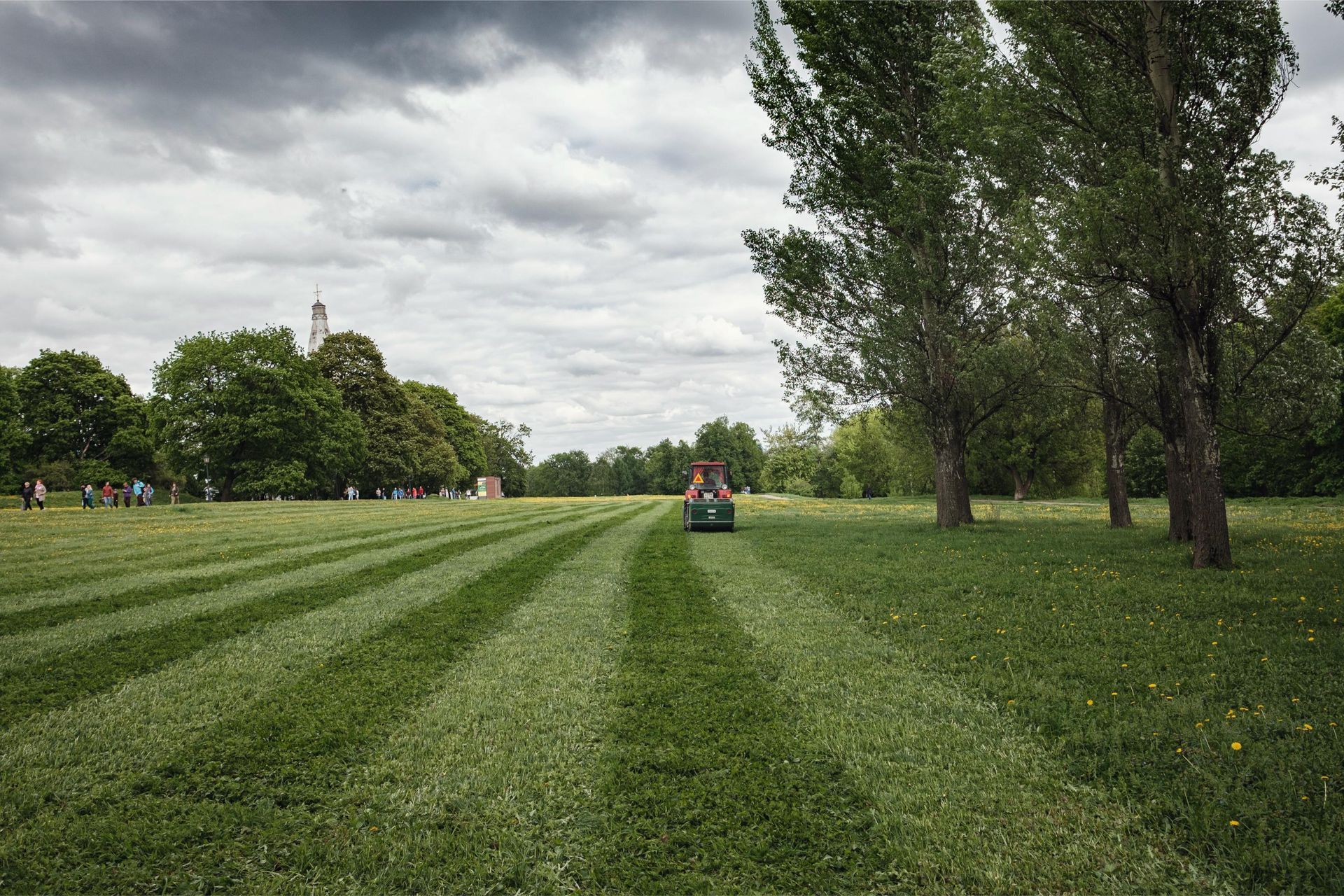 Wide green park lawn with mowed stripes, trees lining the right side, and a small tractor in the distance.