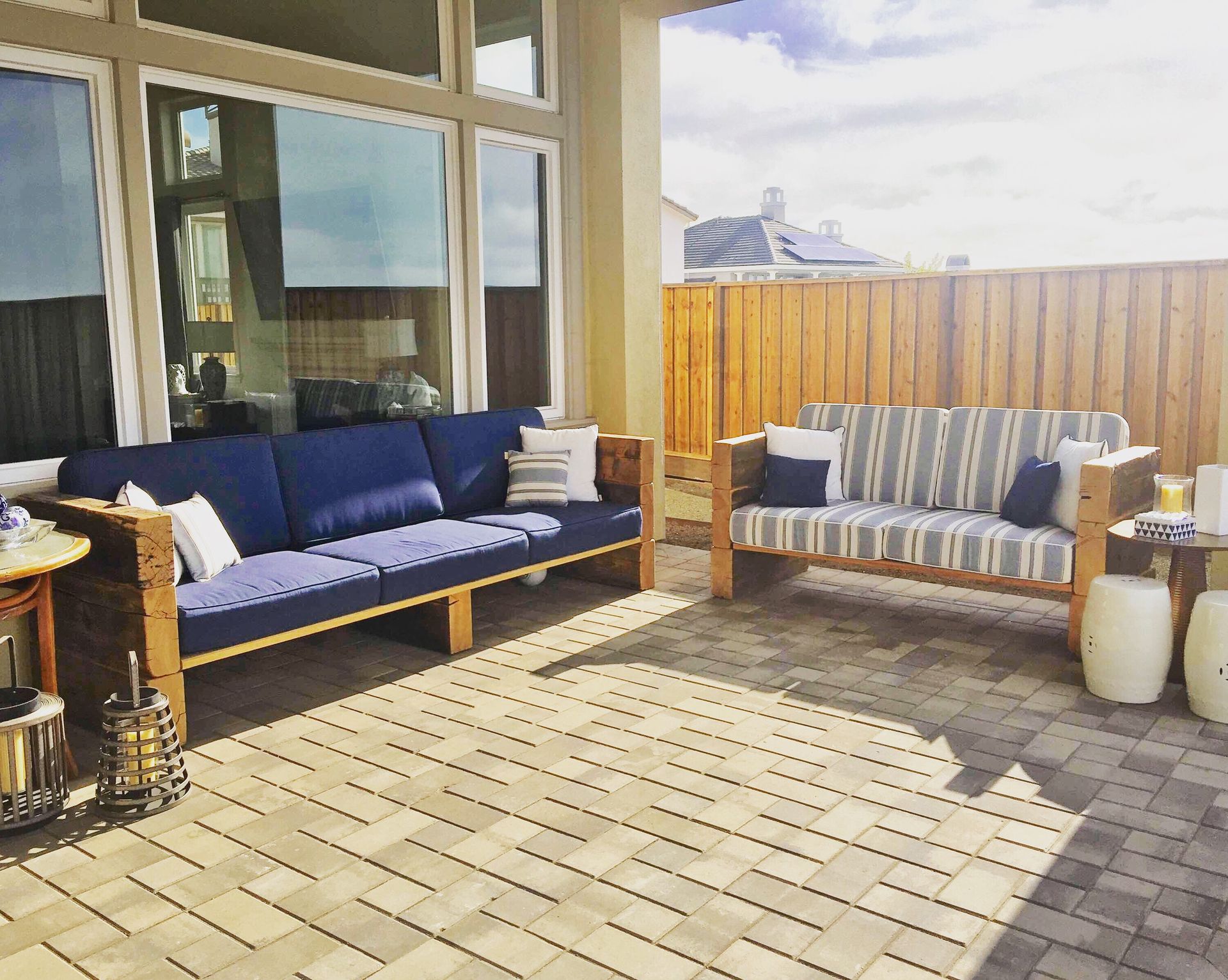Sunlit patio with blue and striped sofas, wooden table, and potted decor beside a house.