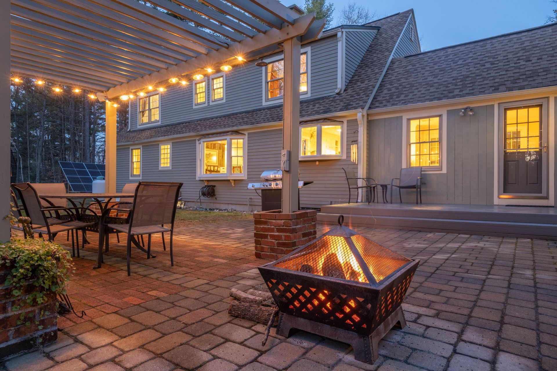 Back patio with fire pit, pergola lights, and outdoor dining set beside a lit house at dusk