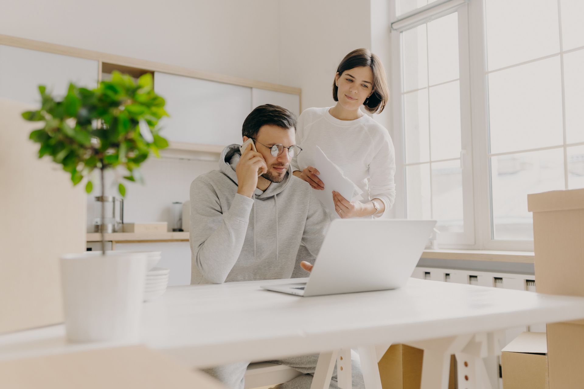 Two coworkers in a bright office, one on a laptop and phone while the other stands beside him.