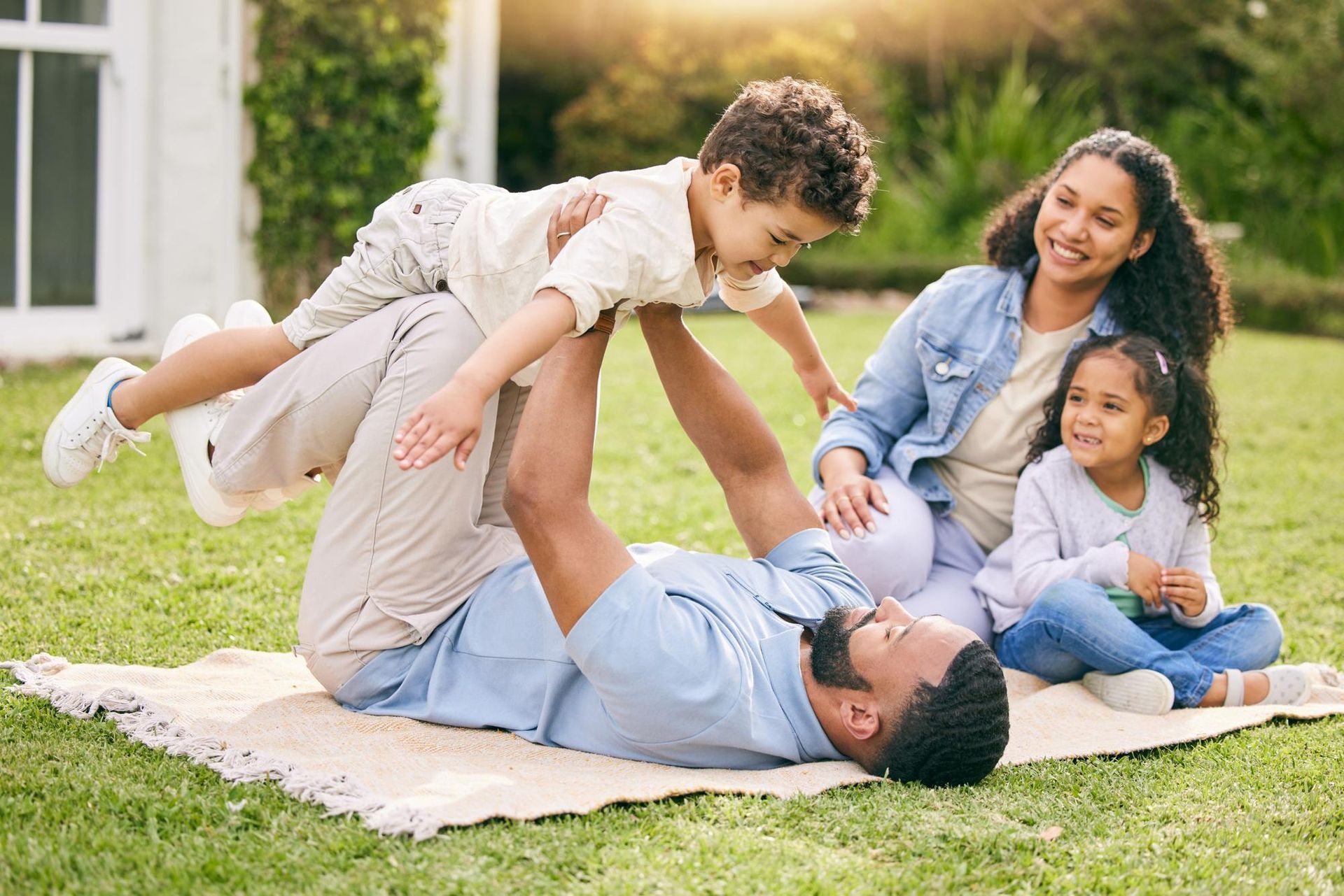 Family on blanket in yard, father lifting child; mother and daughter watch.