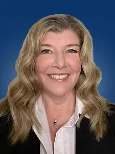 Woman with long blonde hair, smiling, wearing a blazer over a white shirt, against a blue background.