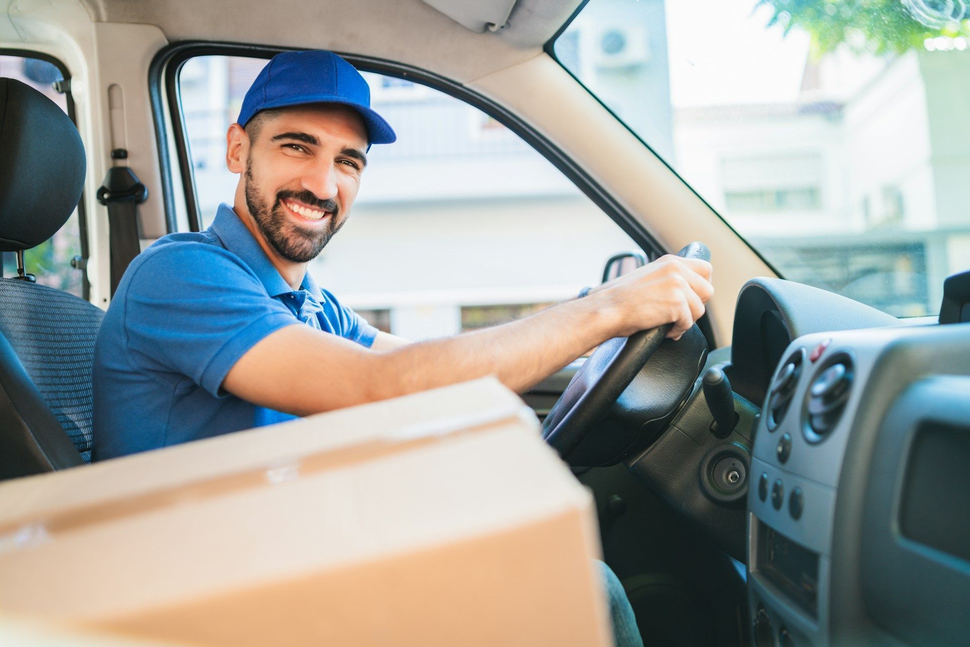 Delivery driver smiling, holding steering wheel inside van, with boxes on the seat.