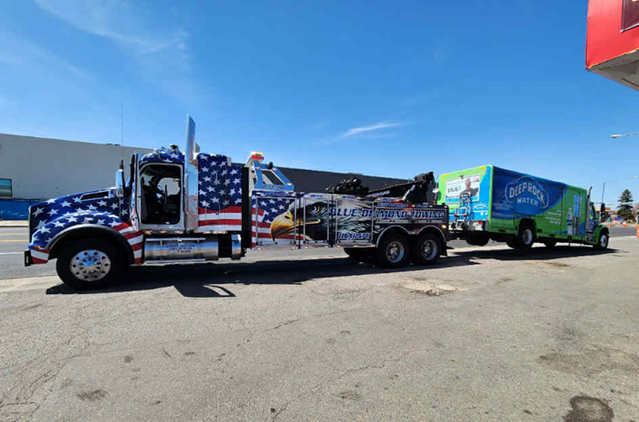 Patriotic tow truck hauling a green and blue box truck on a sunny day.