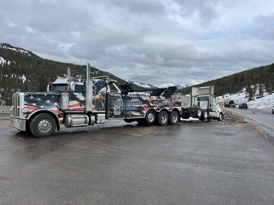 Tow truck hauling a damaged semi-truck on a mountain road with overcast skies.