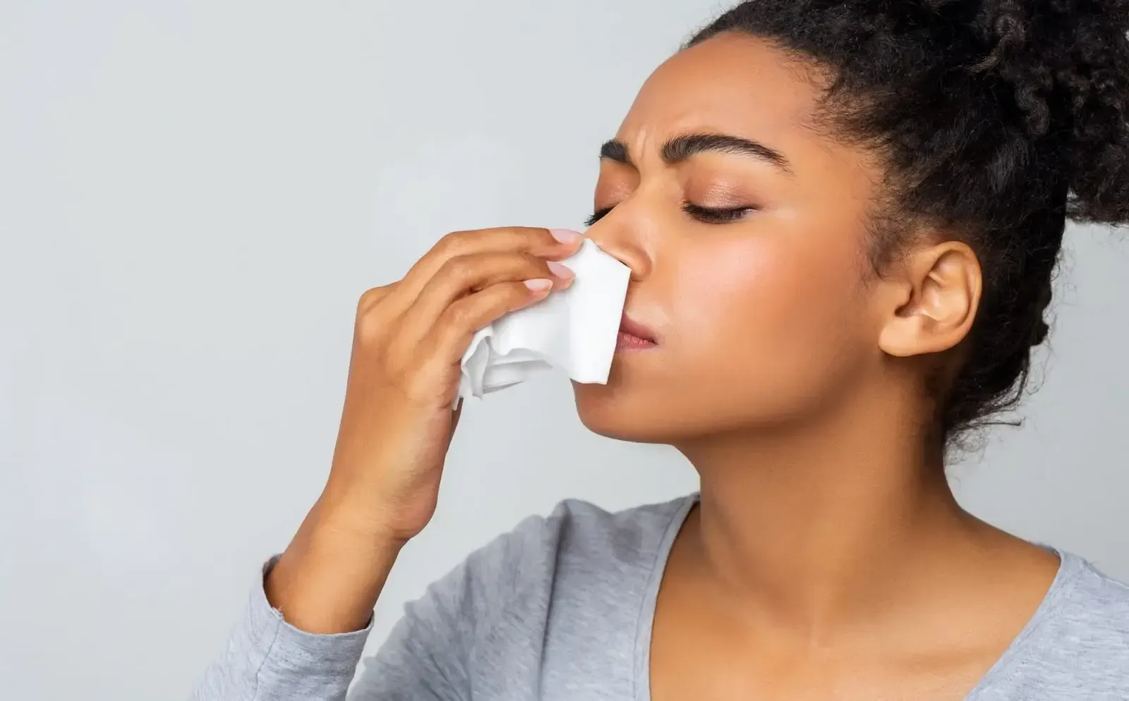 Woman holding tissue to her nose against a light gray background, appearing to blow her stuffy nose