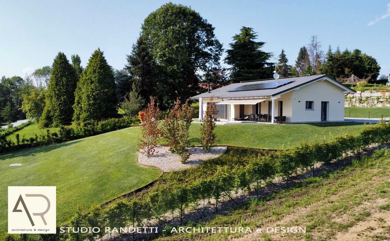 Casa moderna bianca con pannelli solari su un pendio erboso, circondata da alberi sotto un cielo azzurro limpido.
