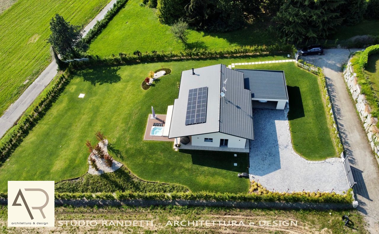 Veduta aerea di una casa moderna con pannelli solari, vialetto di ghiaia e prato verde racchiuso da un muro di pietra.