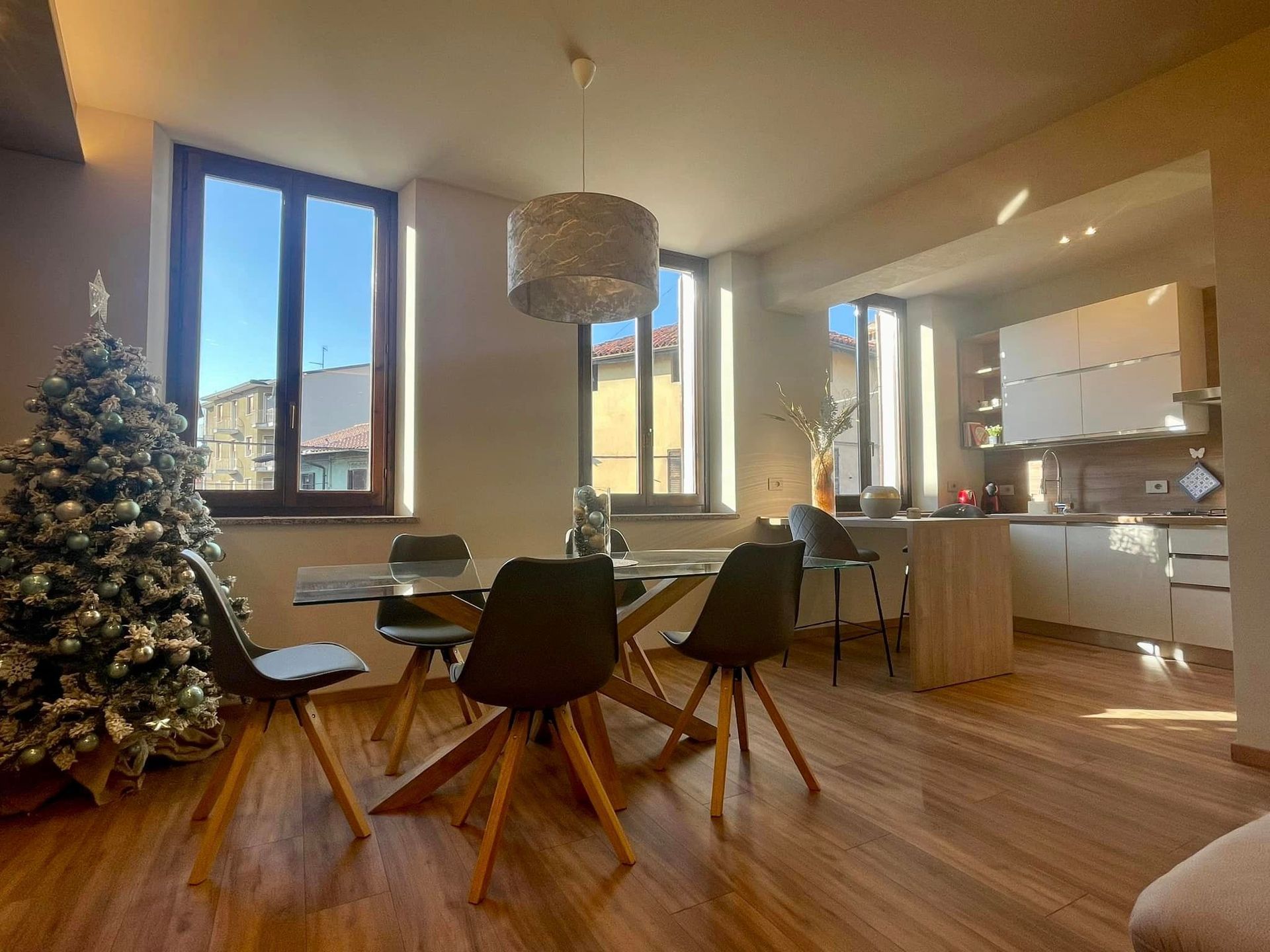Accogliente sala da pranzo con tavolo in legno, sedie nere, albero di Natale e finestre luminose che si affacciano sulla cucina.