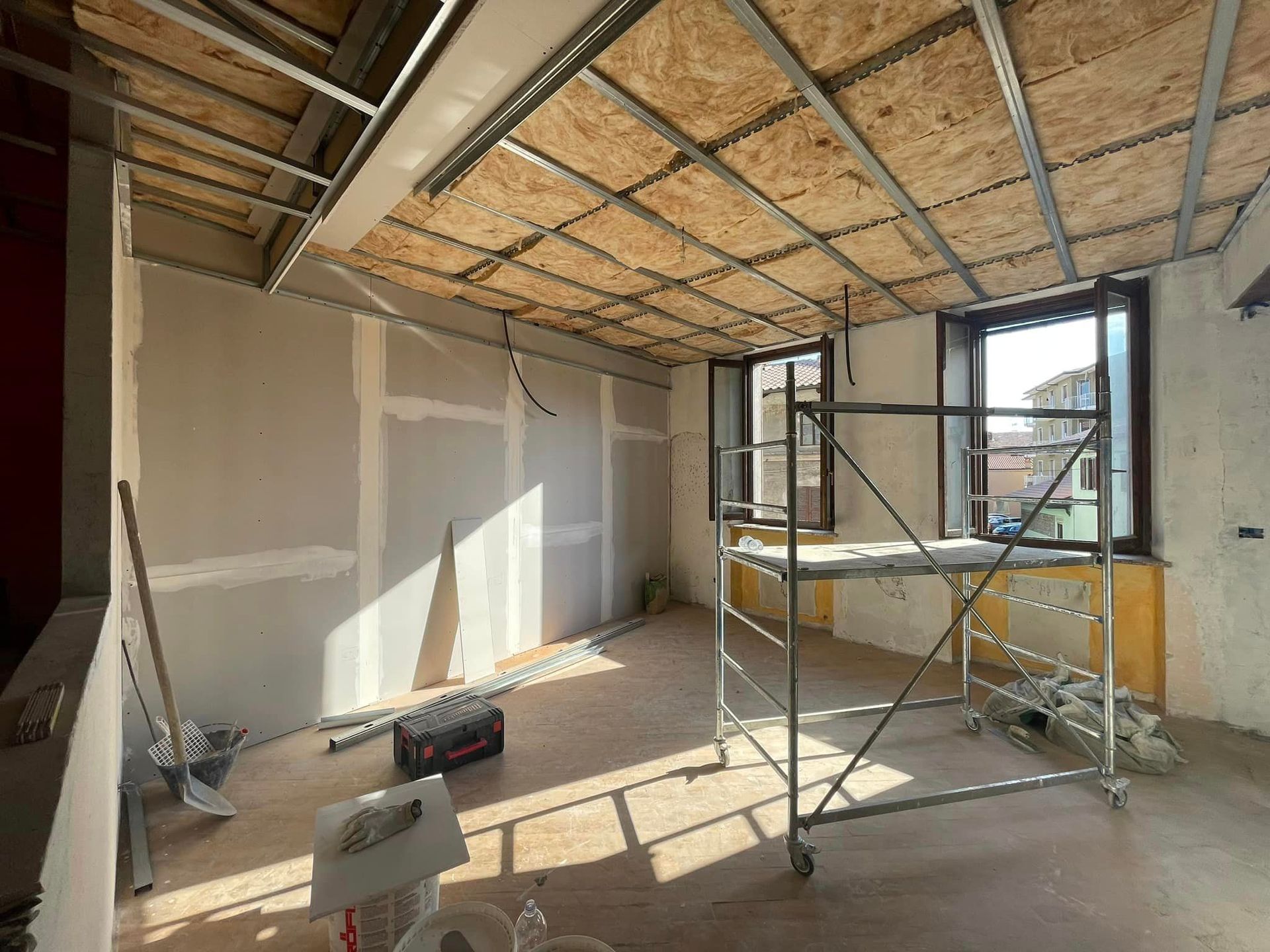 Stanza parzialmente ristrutturata con isolamento del soffitto a vista, impalcature e luce naturale che filtra dalle finestre.