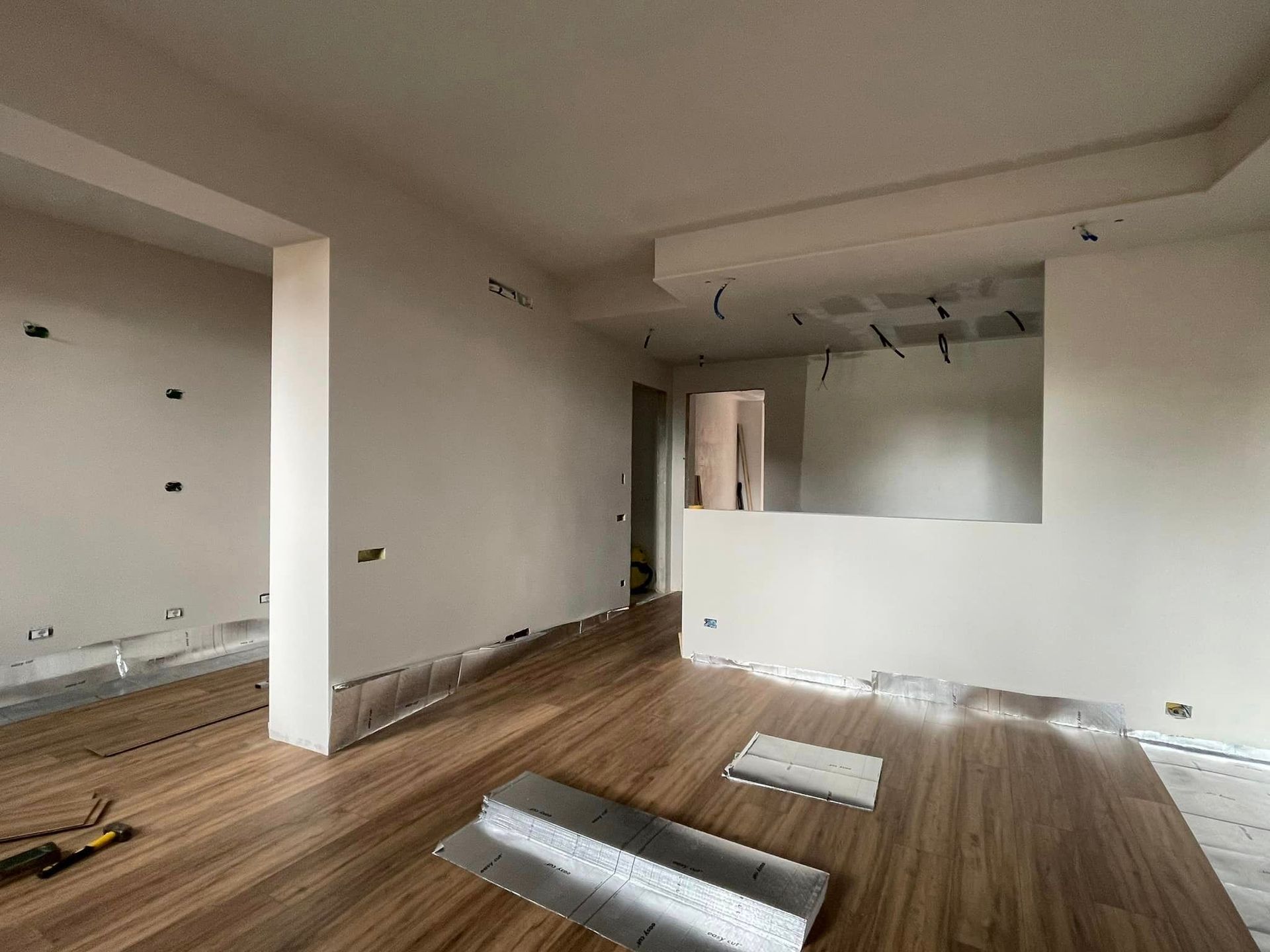 Stanza vuota e non rifinita con pareti bianche, pavimento effetto legno e infissi a vista vicino all'apertura della cucina.