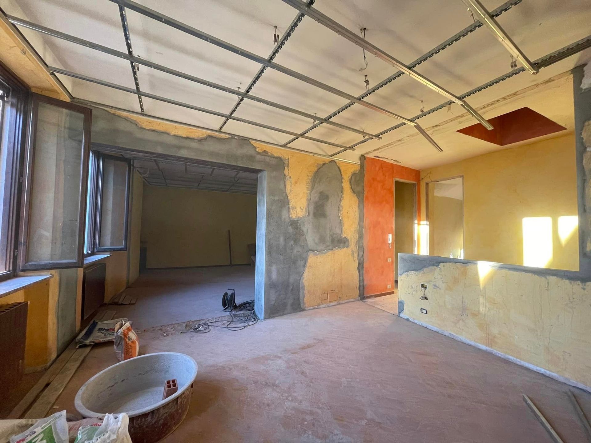 Ristrutturazione in corso in una stanza non rifinita con soffitto a vista, pareti in intonaco e detriti di costruzione.