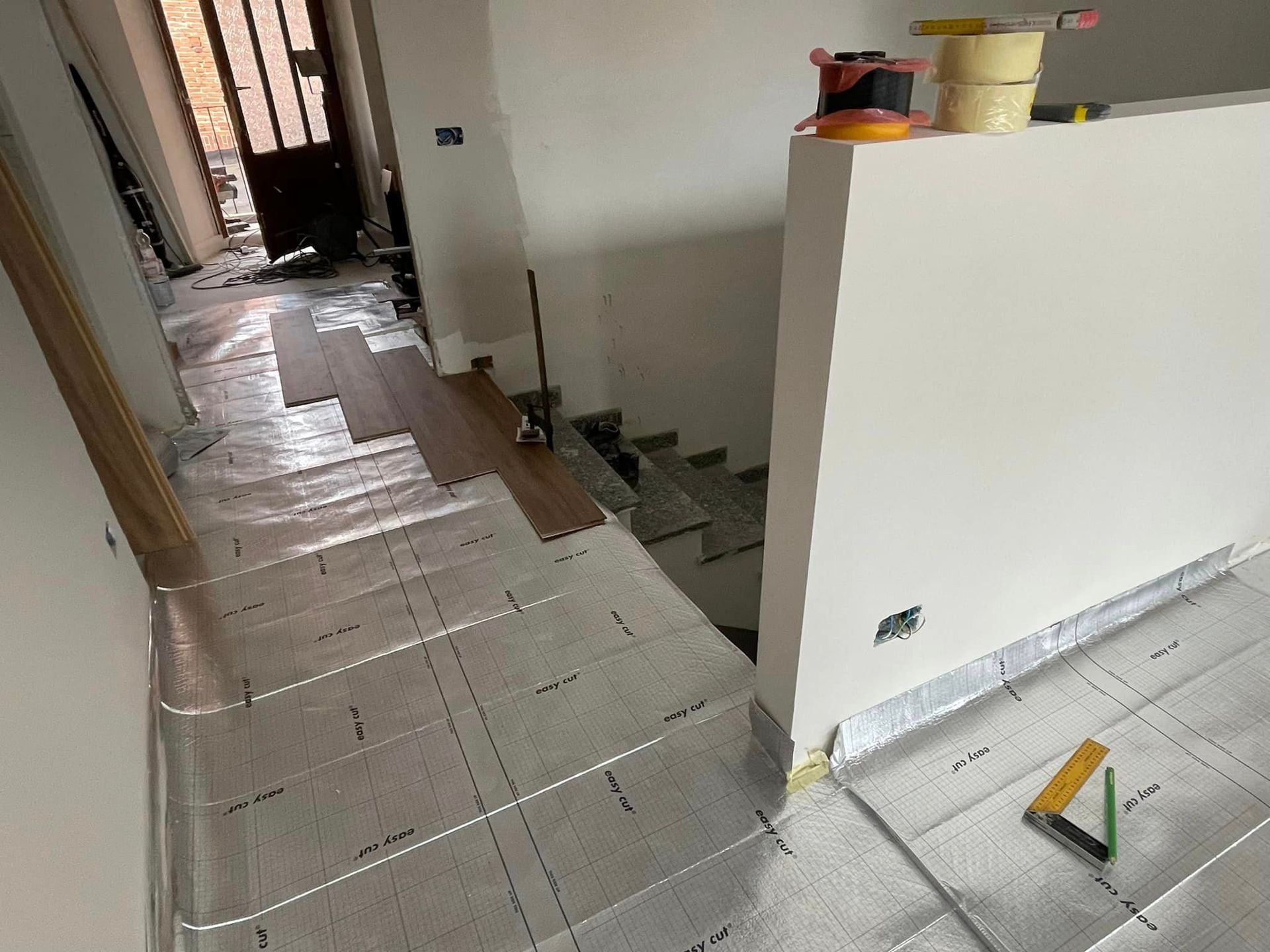 Stretto corridoio in fase di ristrutturazione con pavimento in piastrelle bianche, parziale parete divisoria e attrezzi sulla destra.