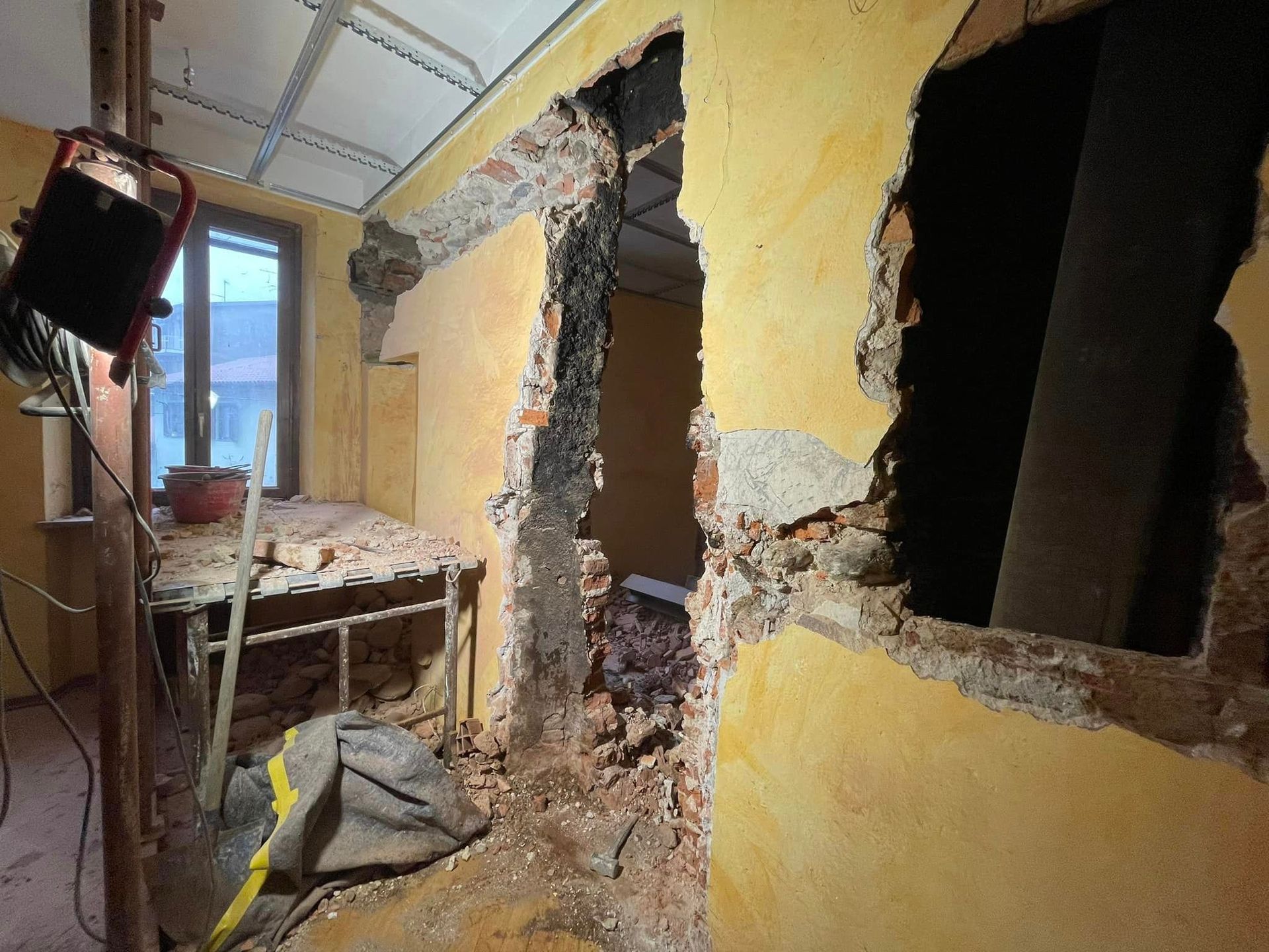 Corridoio gravemente danneggiato con pareti gialle rotte, macerie a vista e detriti vicino a una finestra.