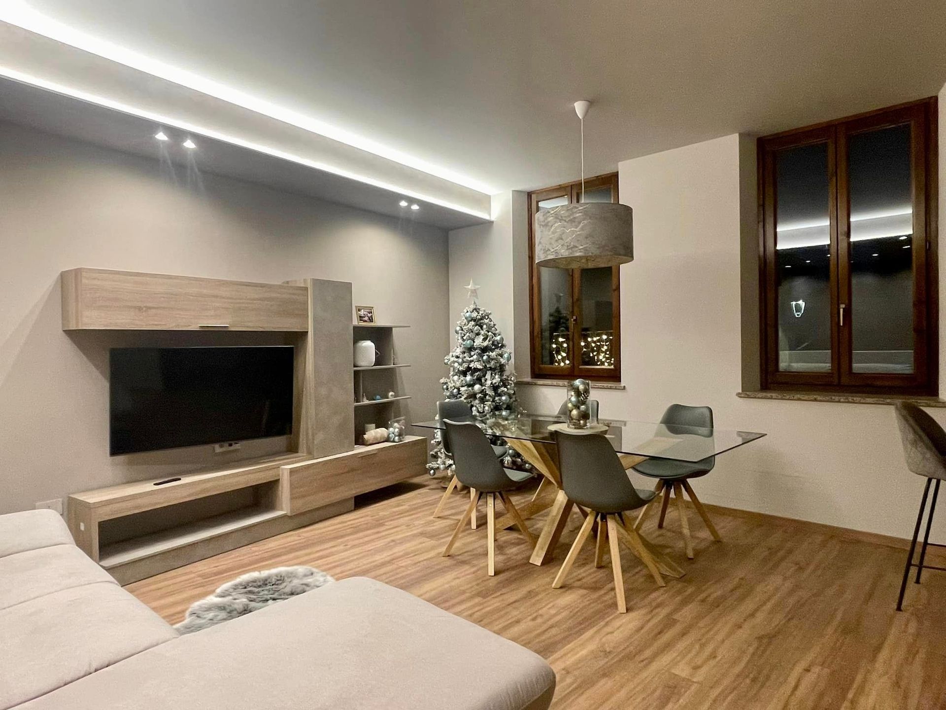 Soggiorno moderno con TV a parete, albero di Natale, pavimento in legno e tavolo da pranzo vicino a una finestra.