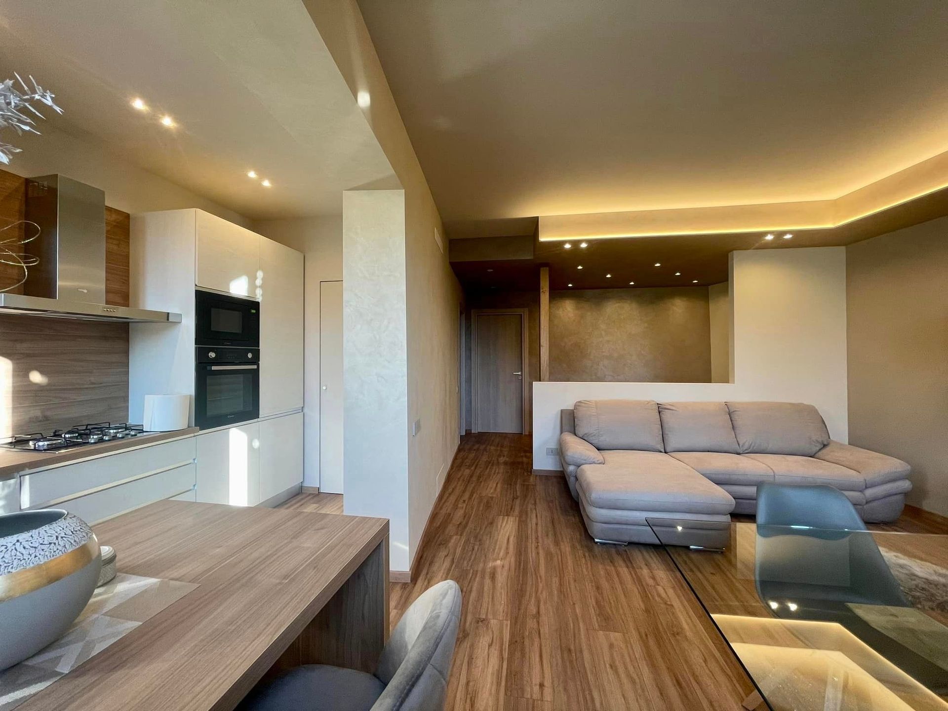 Soggiorno moderno a pianta aperta con divano angolare grigio, pavimento in legno e illuminazione a incasso dai toni caldi.