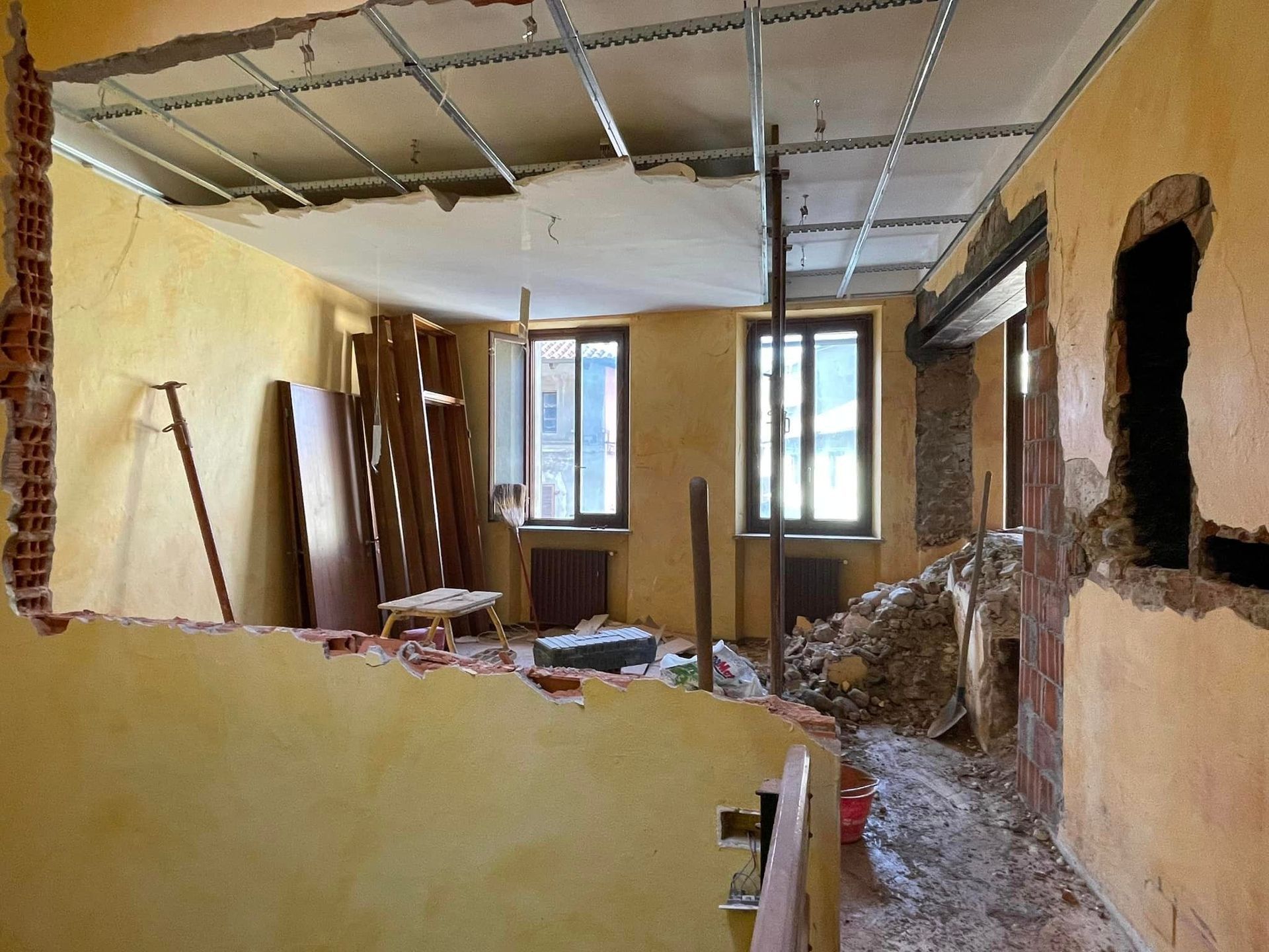 Stanza danneggiata con travi del soffitto a vista, detriti sul pavimento e finestre lungo la parete di fondo.