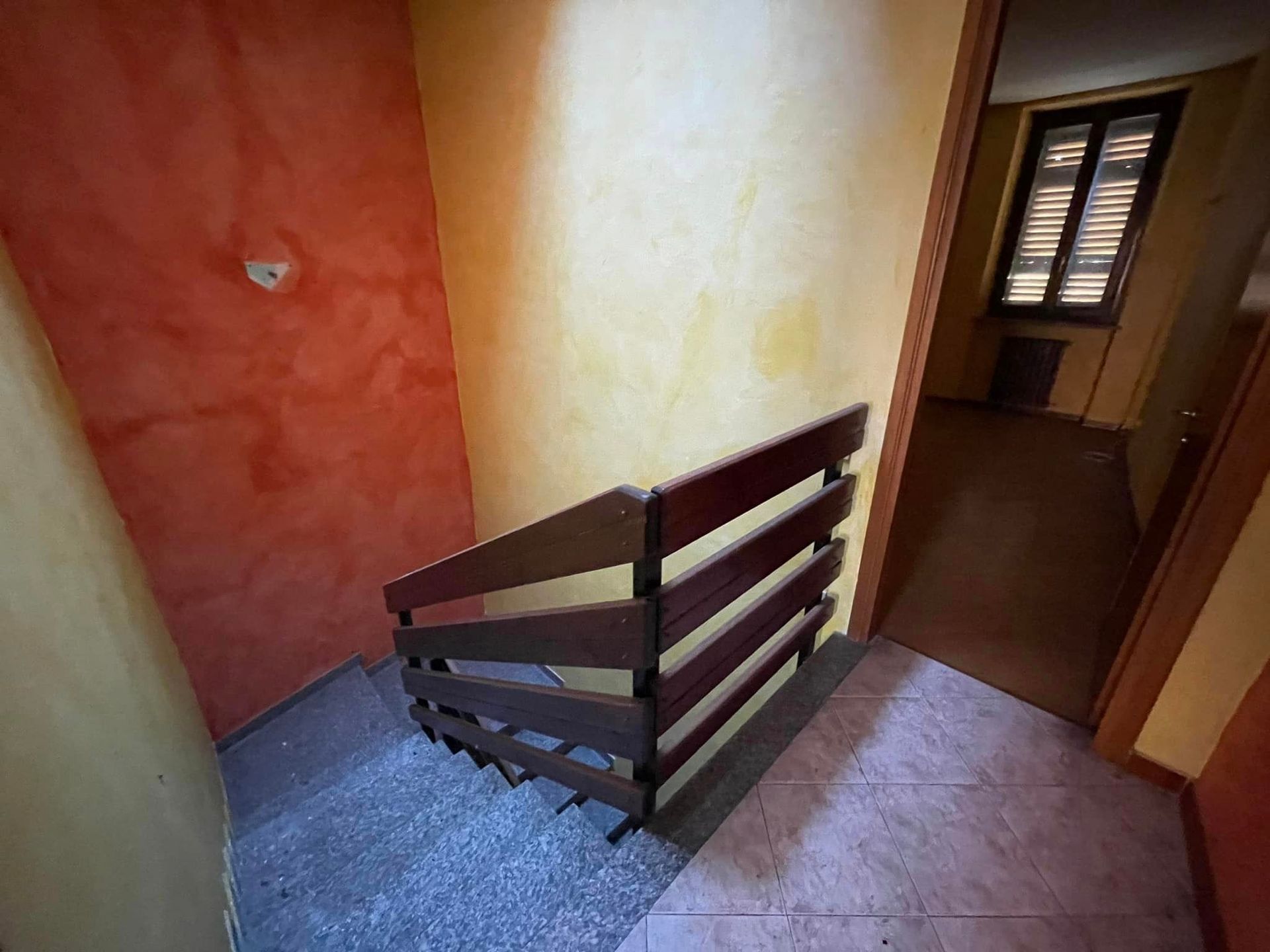 Pianerottolo di una scala interna con ringhiera in legno tra pareti arancioni e color crema, che conduce a una porta e a una finestra.