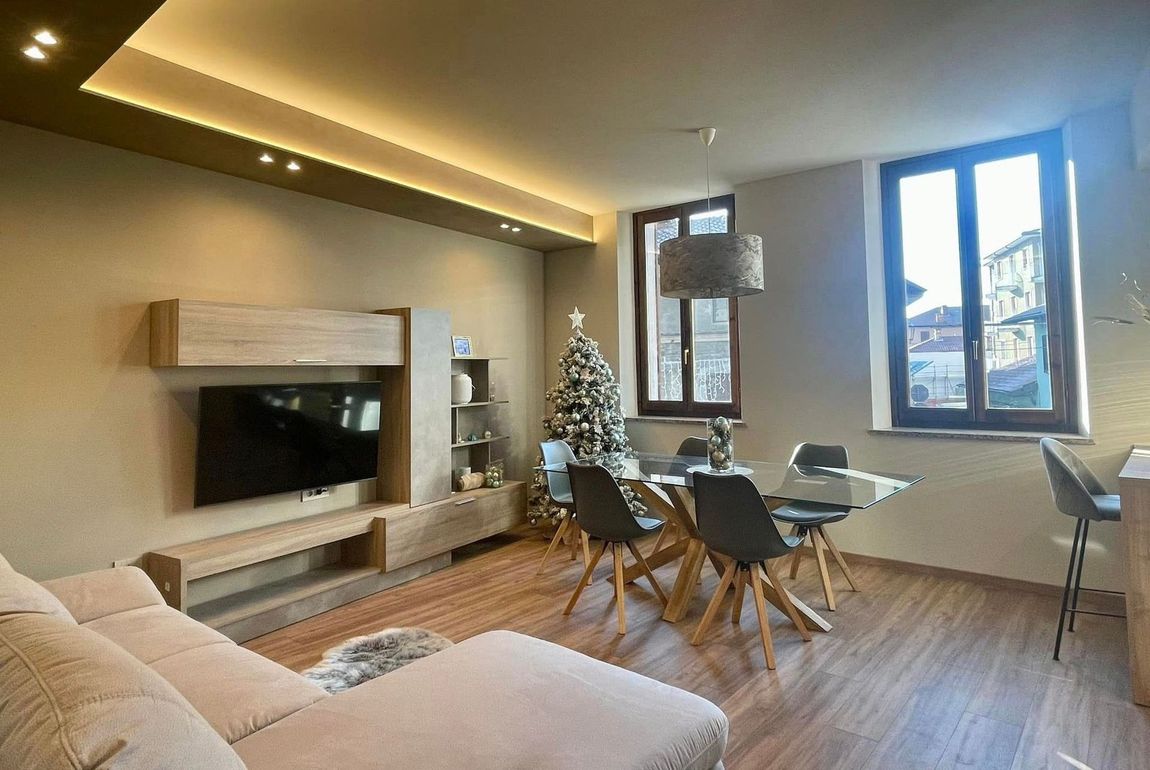 Soggiorno moderno con divano beige, TV a parete, tavolo da pranzo e ampie finestre con vista sulla città.