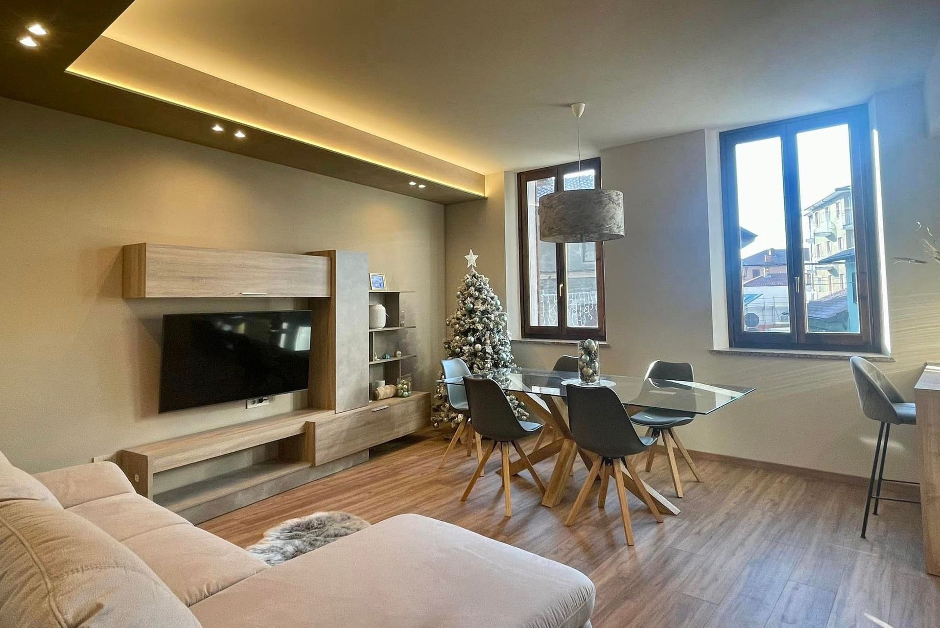 Soggiorno moderno con divano beige, parete attrezzata con TV, tavolo da pranzo e ampie finestre con vista sulla città.