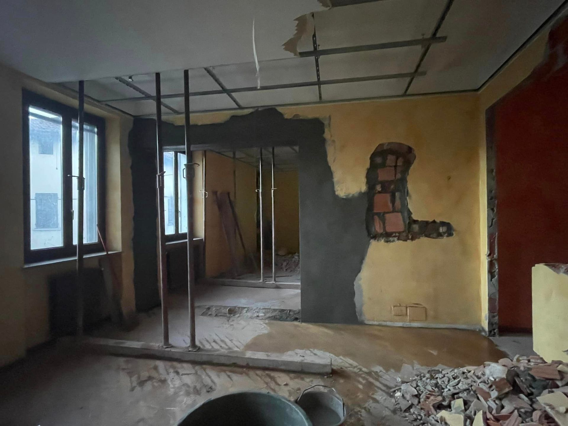 Stanza parzialmente ristrutturata con travi a vista, detriti sul pavimento e una finestra sulla sinistra.