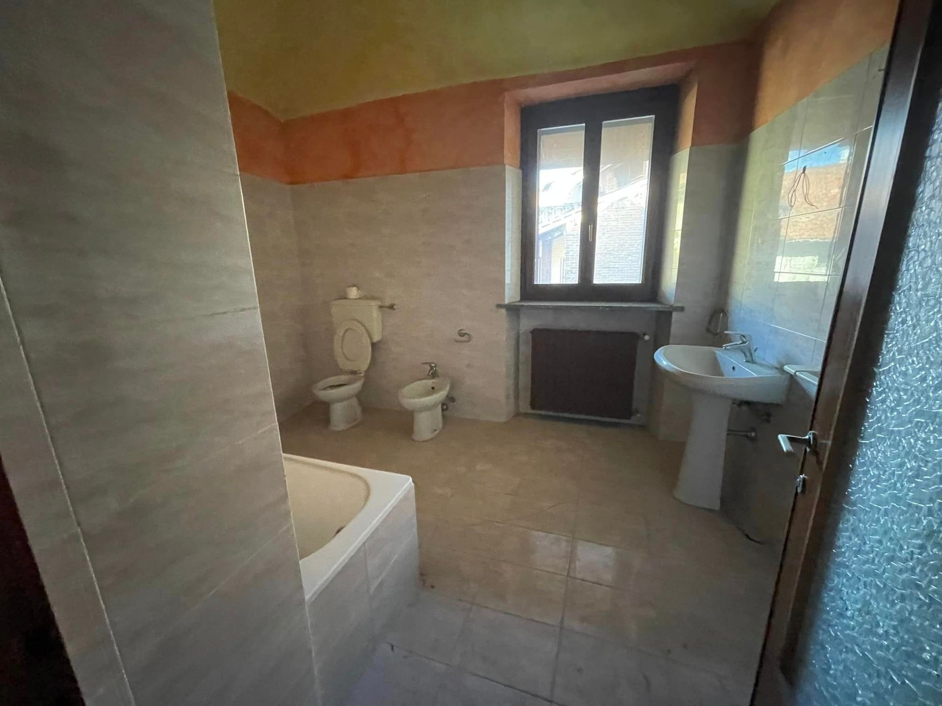 Piccolo bagno con vasca, WC, bidet, lavabo e finestra, pareti e pavimento piastrellati beige.