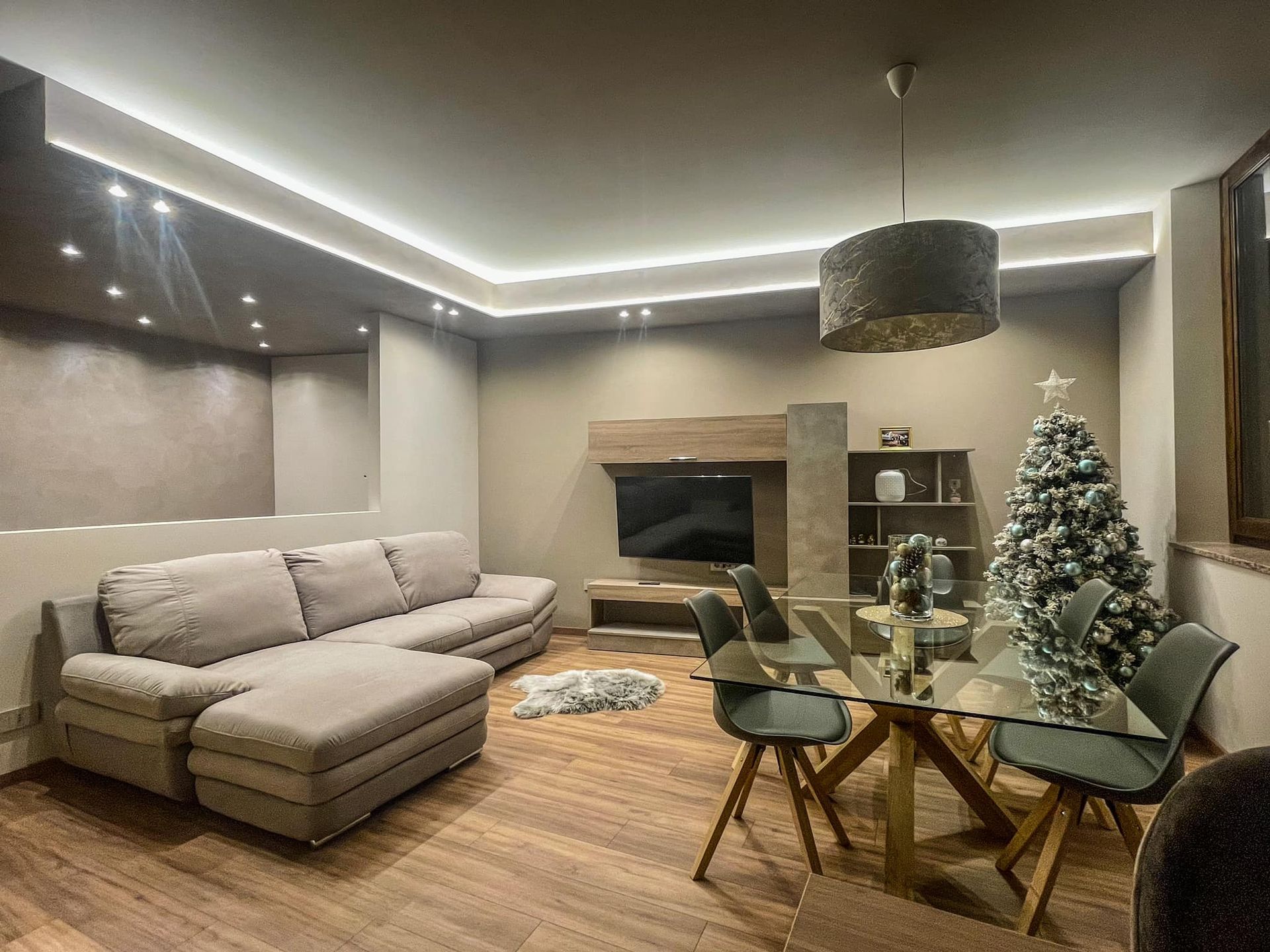 Soggiorno moderno con divano componibile grigio, tavolo da pranzo in vetro, camino e albero di Natale decorato.