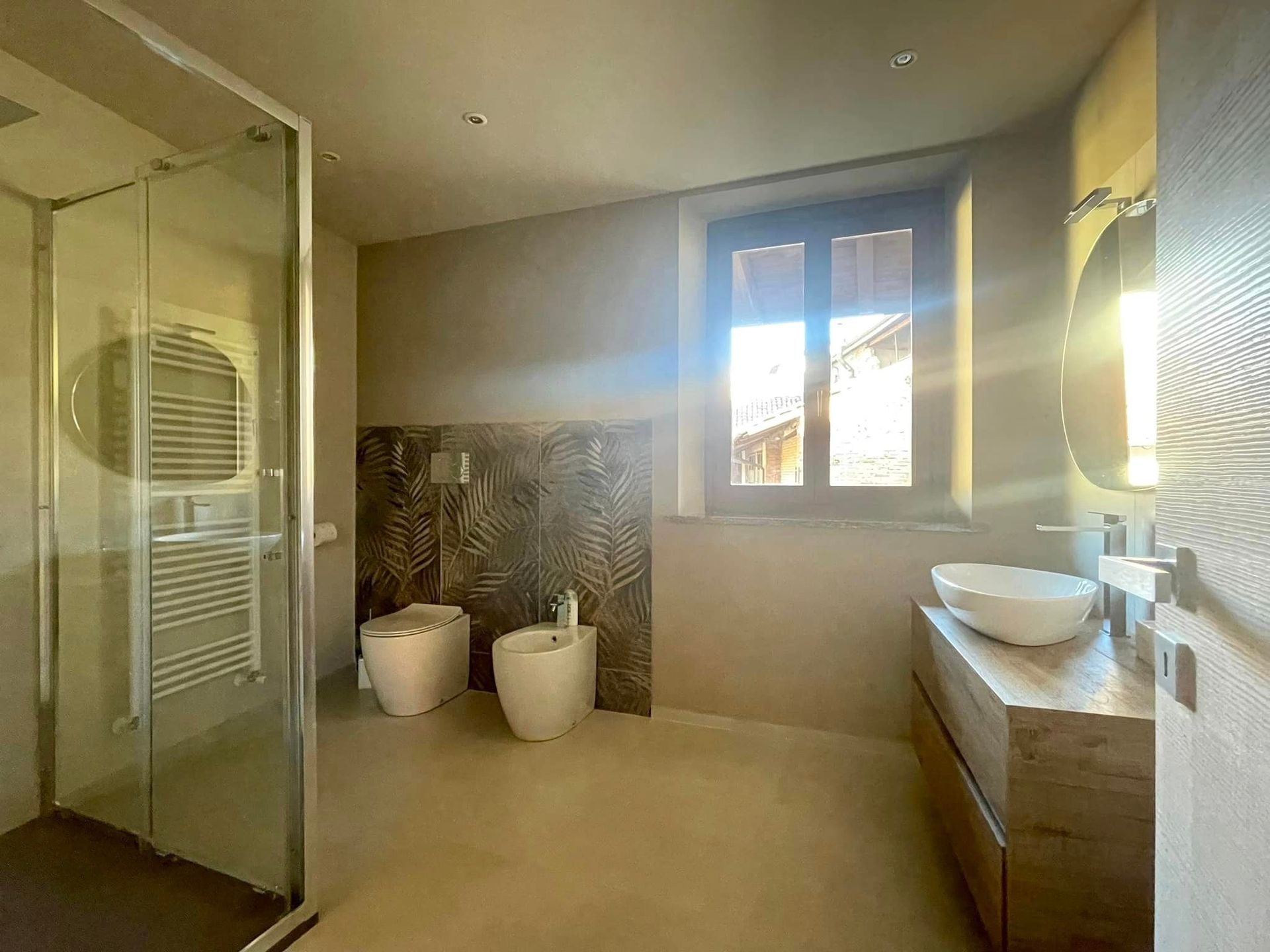 Bagno moderno color beige con doccia in vetro, vasca freestanding e luminosa finestra.